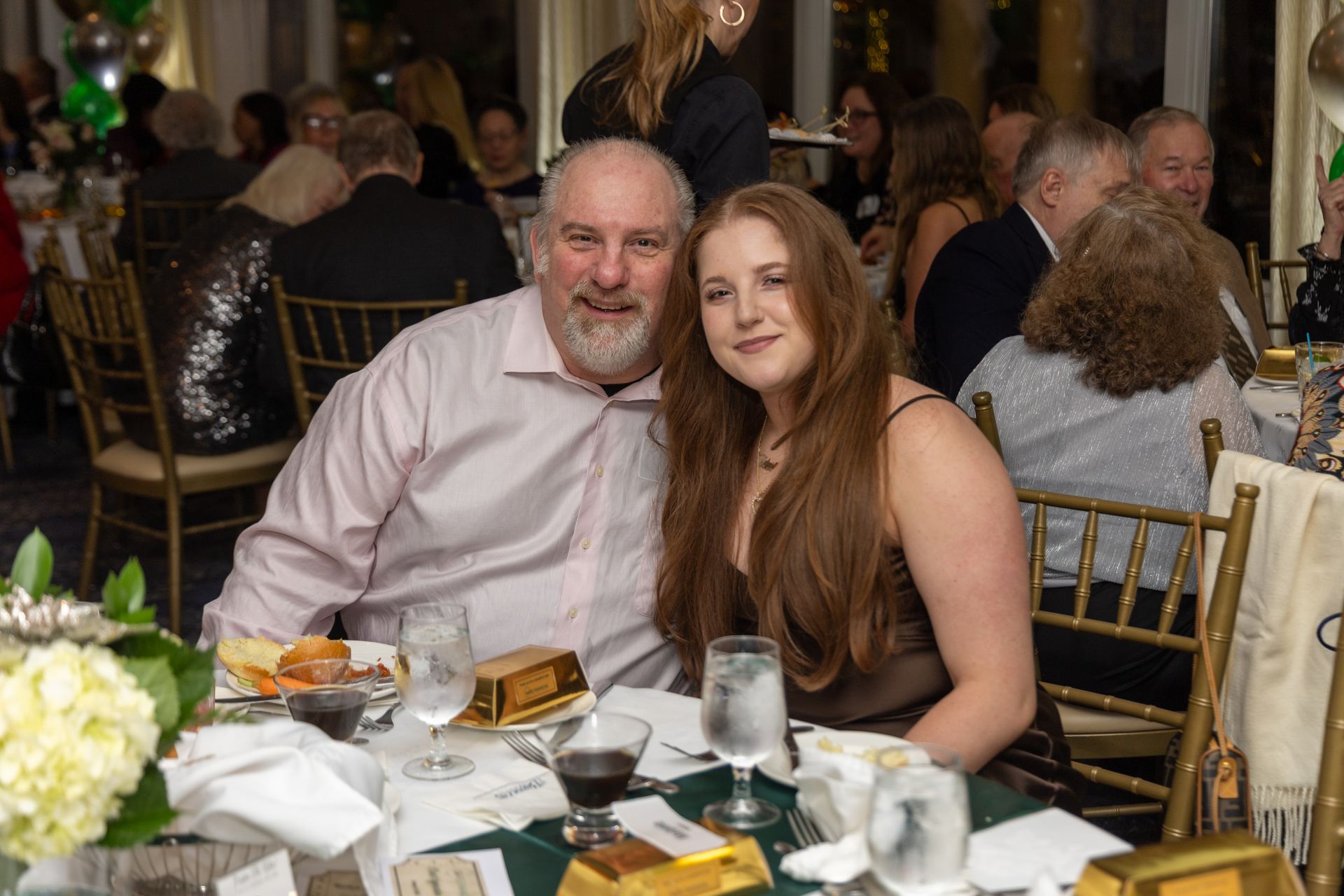Man and woman at a gala, smiling. They sit at a table with food, drinks, and decor.