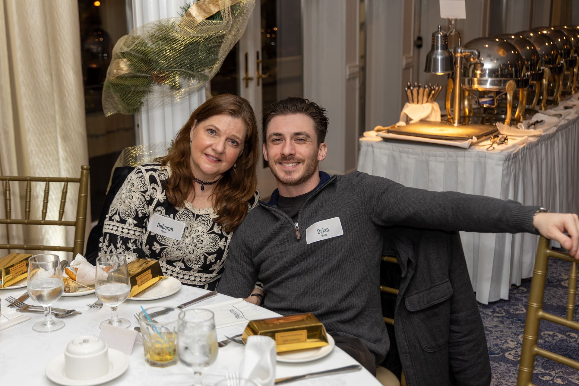 Two people smiling at a table set for a meal, a buffet in the background.