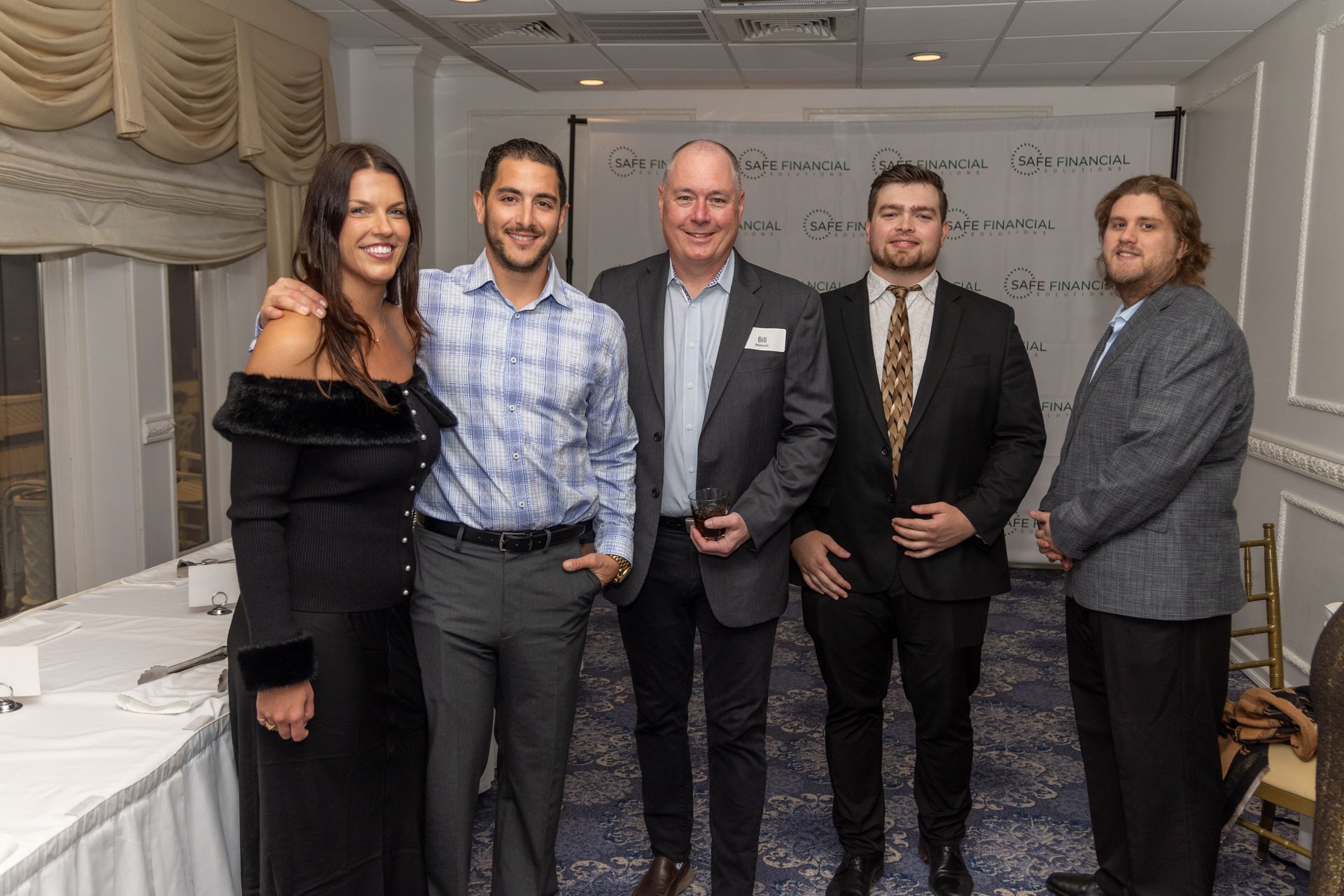 Five people posing for a photo indoors, a woman in a black dress, four men in suits, smiling.