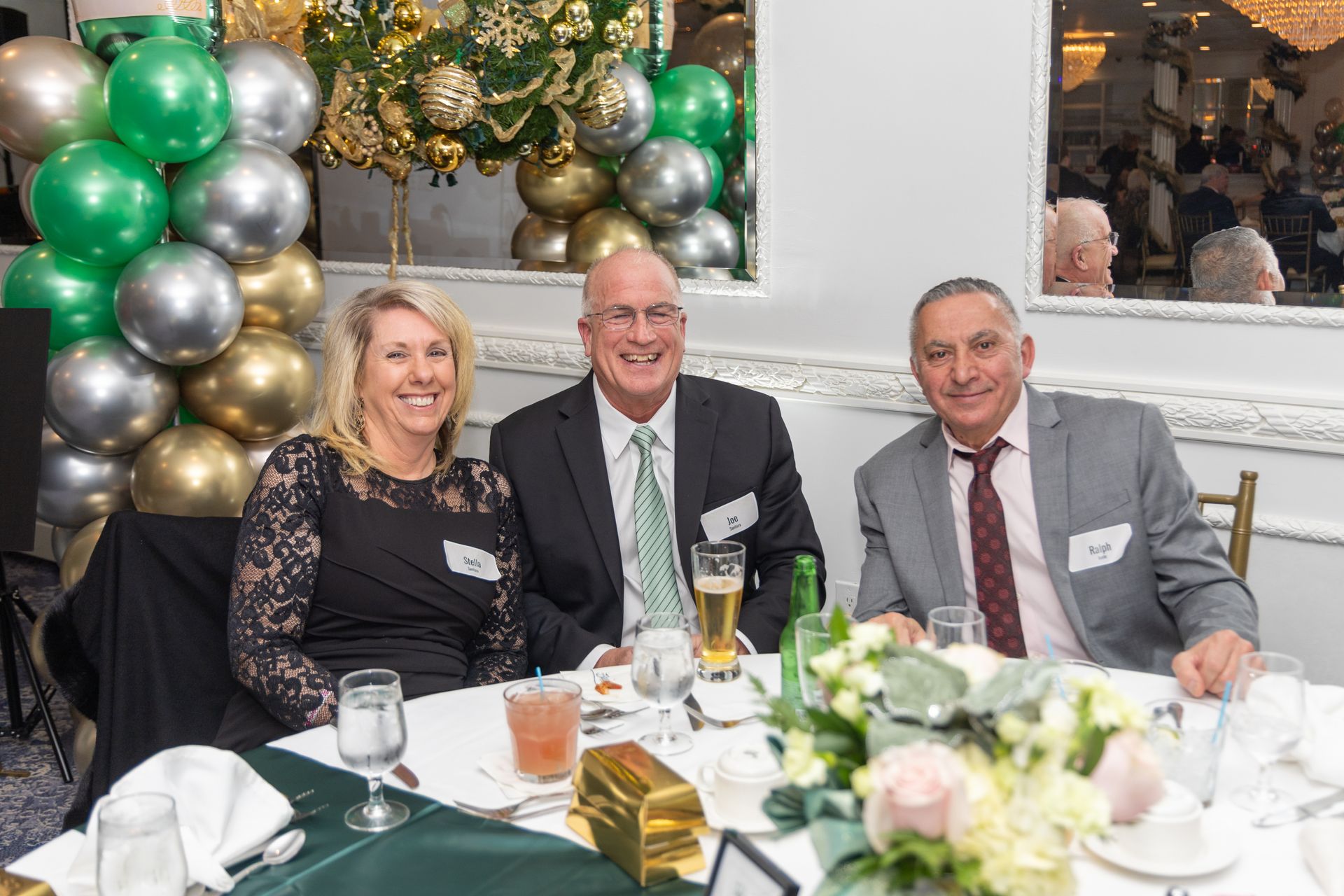 Three people smiling at a table, likely a formal event. Silver, gold, and green balloons in background.
