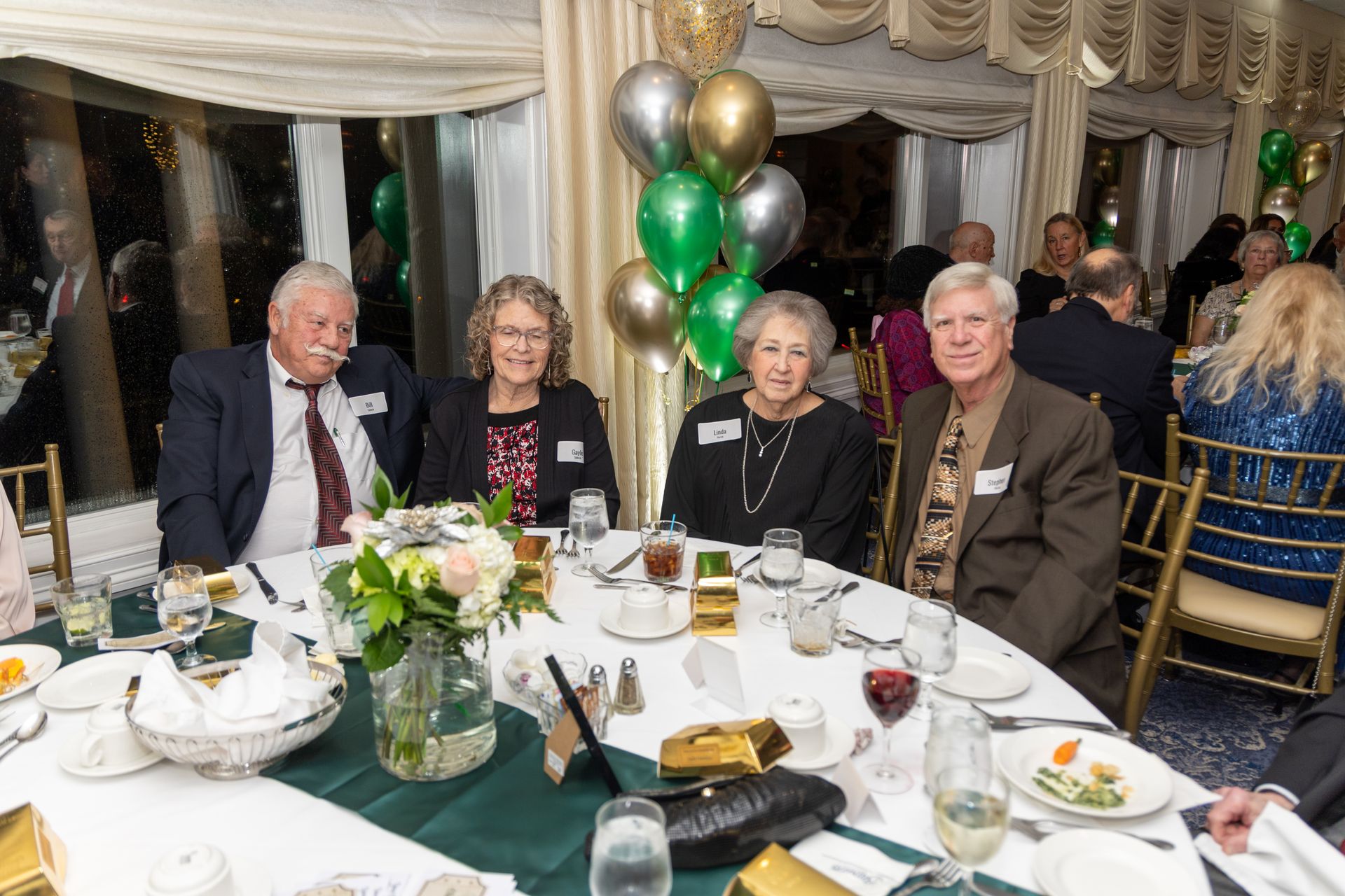 Four people seated at a table in a banquet hall, celebrating. Balloons in the background.