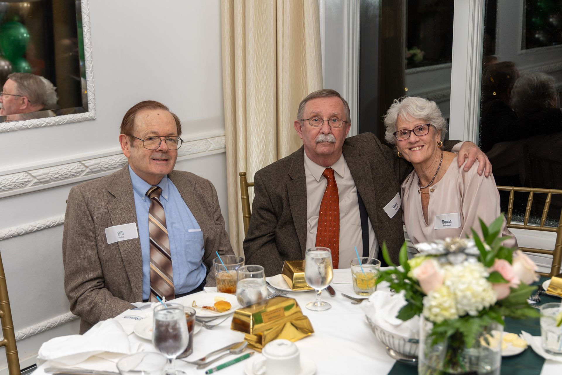 Three people at a table, smiling. Man in the middle has arm around woman. Table set with flowers, glasses.