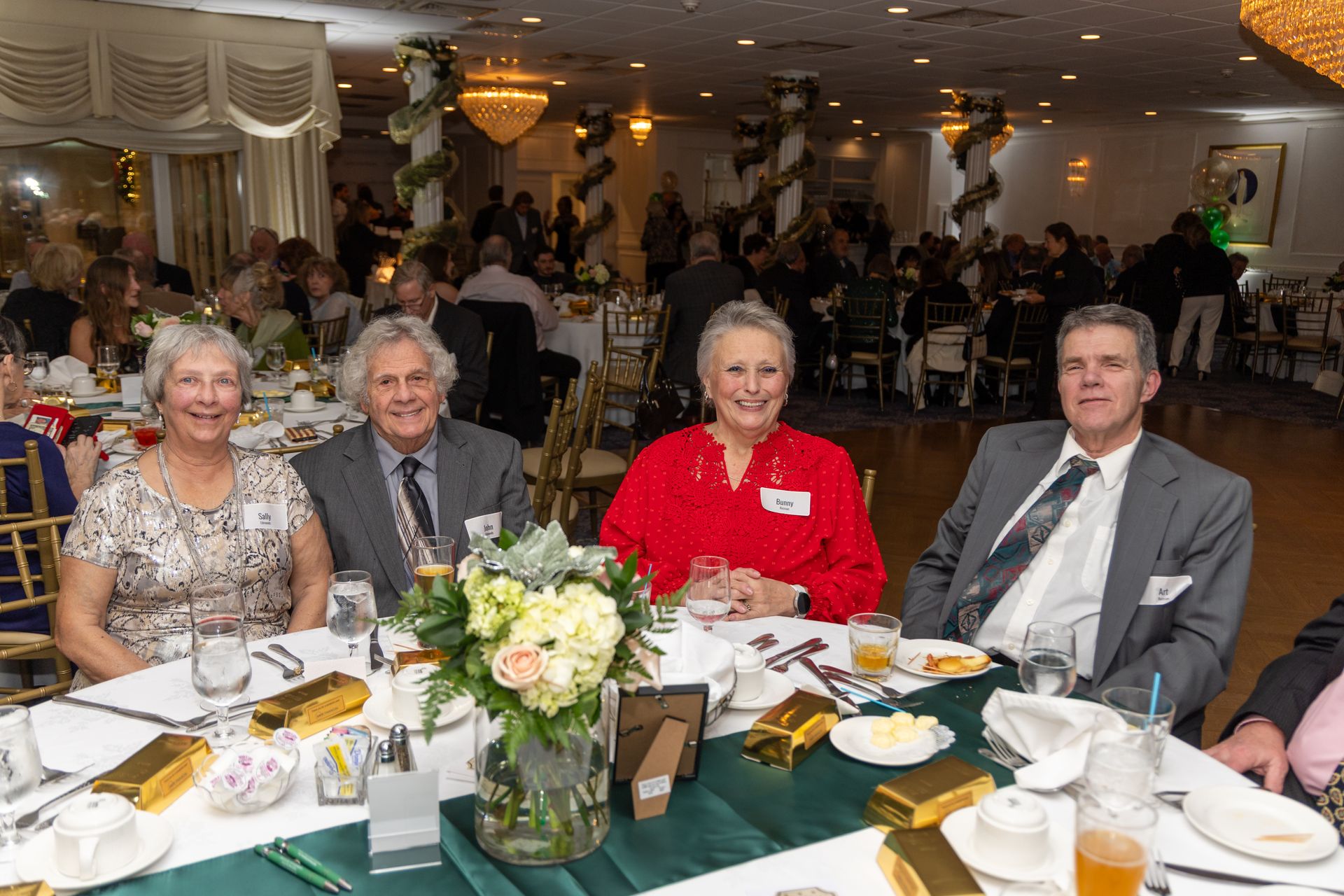 Four people seated at a table in a banquet hall, smiling. A flower arrangement and place settings are visible.