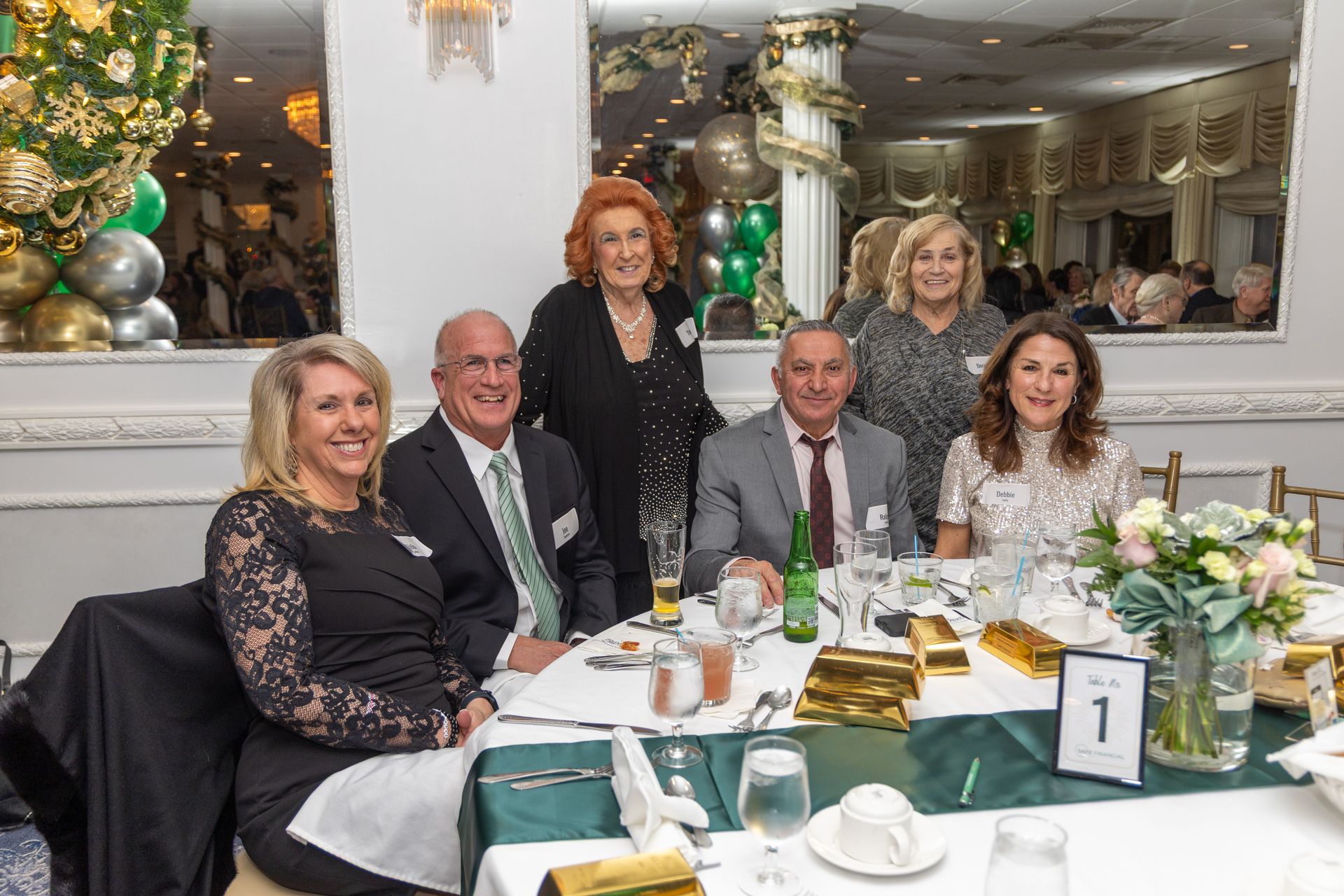 Group of people at a formal dinner, smiling around a table set with gold and green accents, decor.