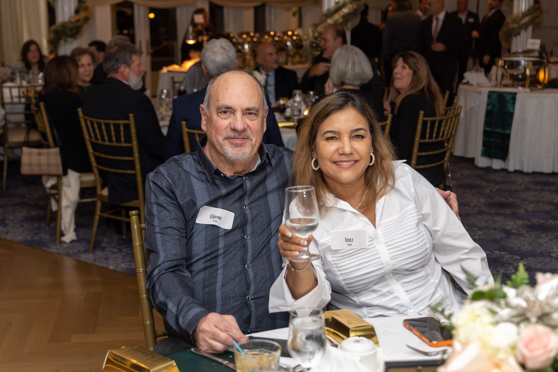 Couple at a formal event; woman raises a glass. They are at a table, smiling, with others in the background.