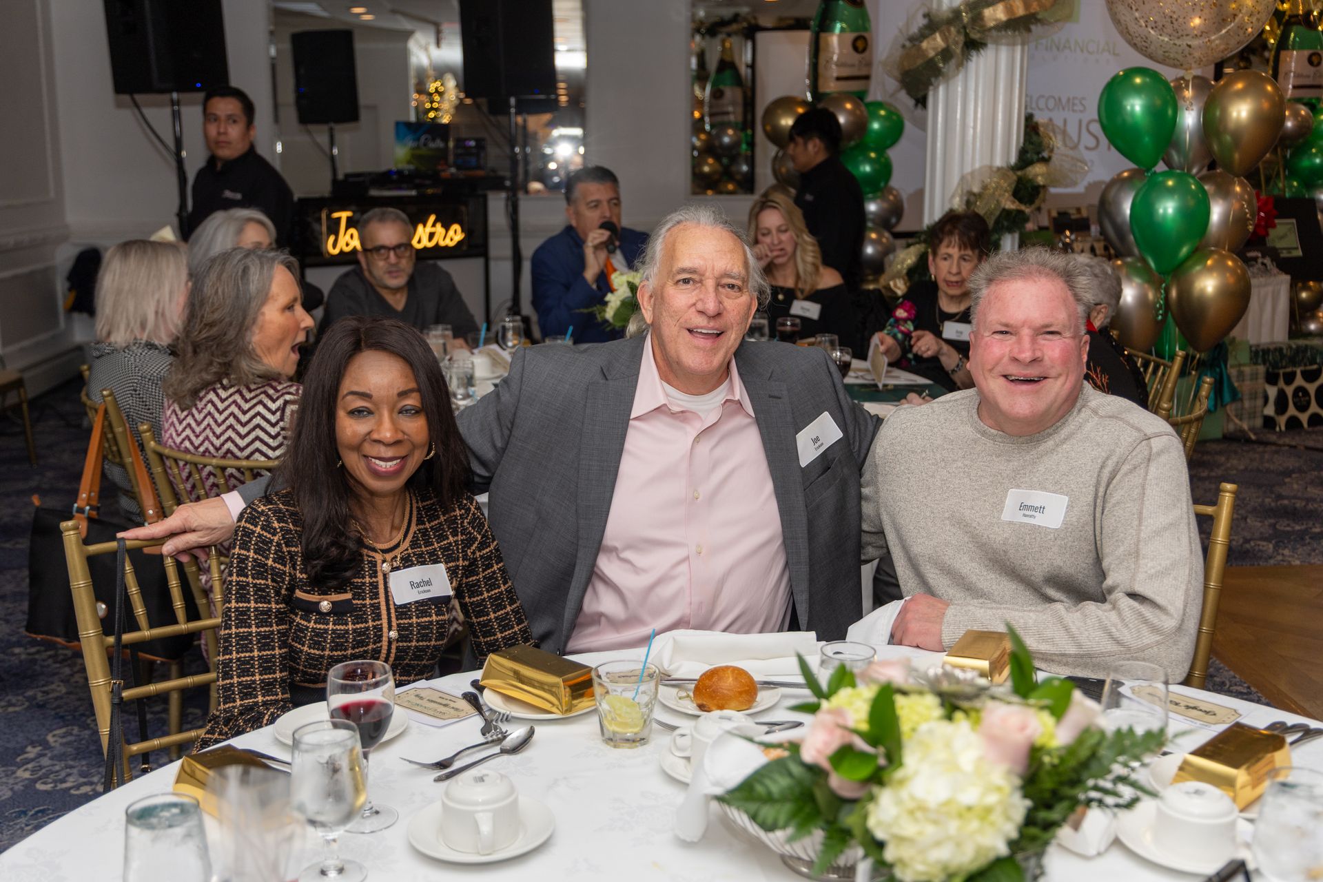 Guests seated at a decorated table, smiling at a social gathering. Balloons and flowers visible in a banquet hall.
