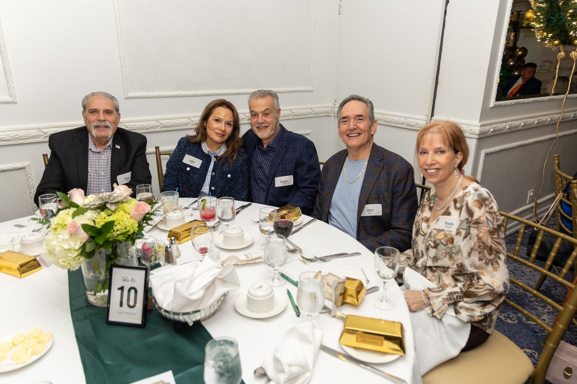 Five people seated around a table at a formal event, smiling. Floral arrangement and place settings are visible.