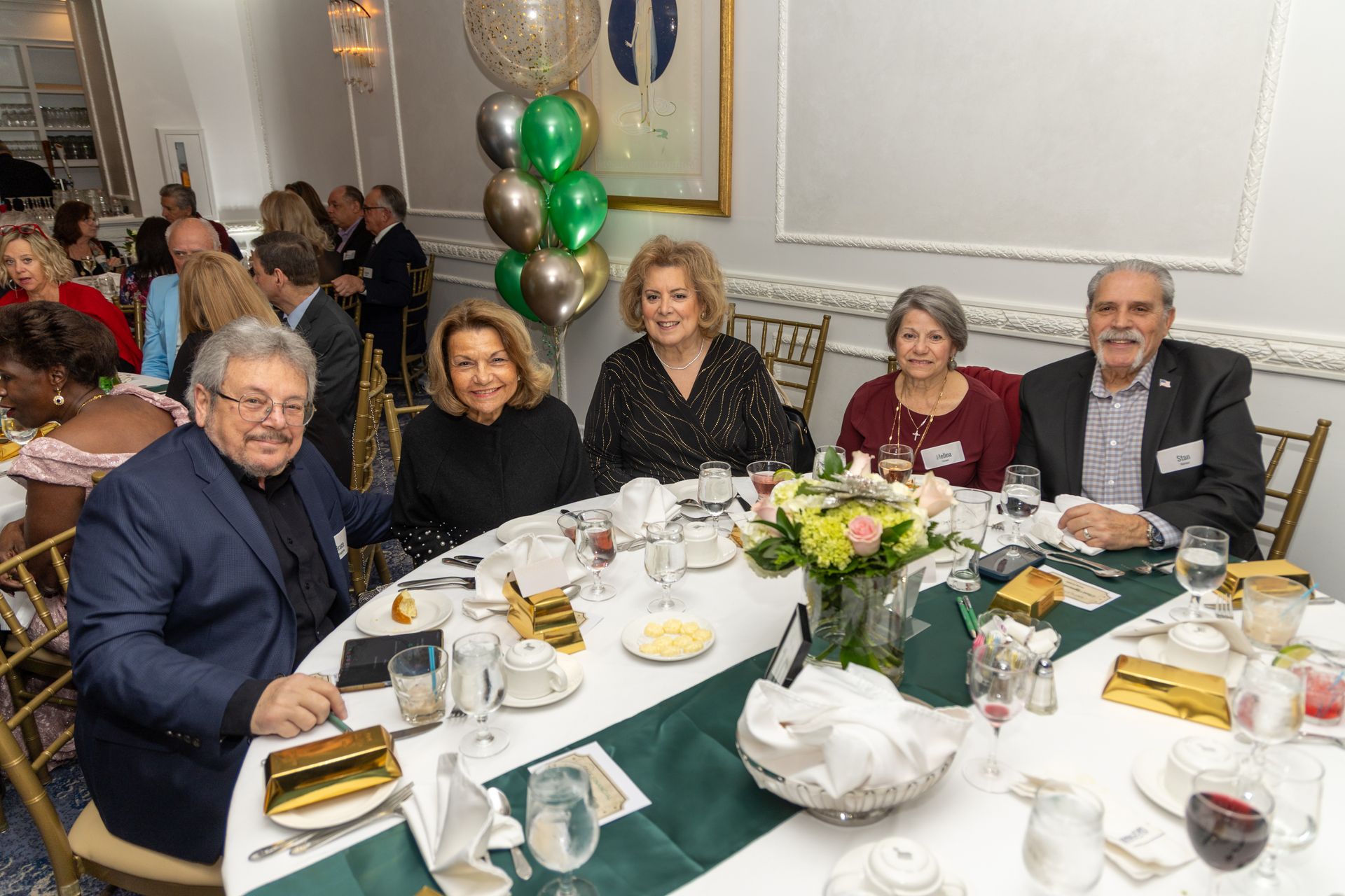 People seated at a decorated table, likely a luncheon. Balloons behind, floral centerpiece, gold accents.