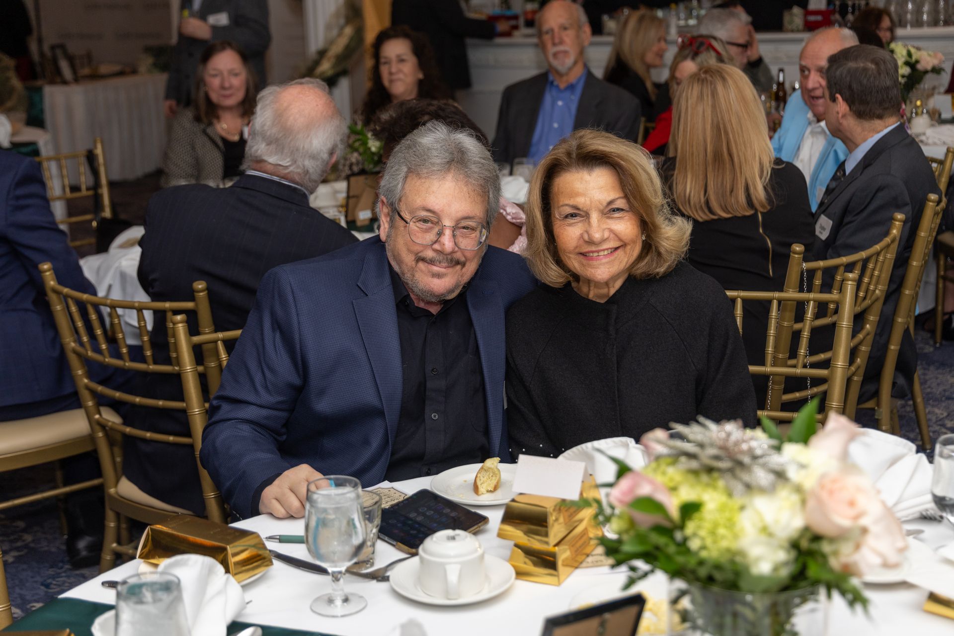 Couple smiling at a table set for a formal event, with flowers and guests in the background.