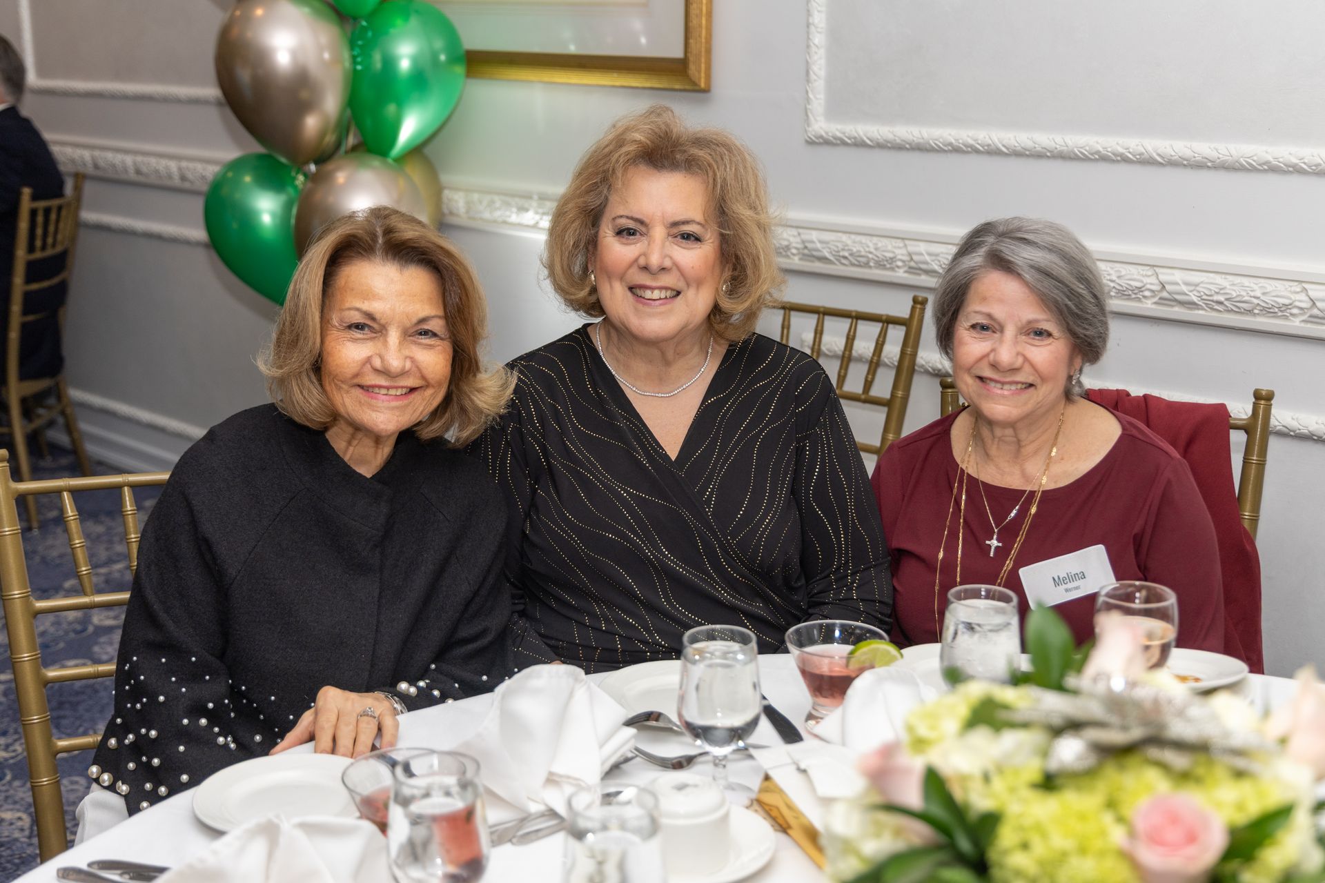 Three women smile at a table set for a meal, gold and green balloons in the background.