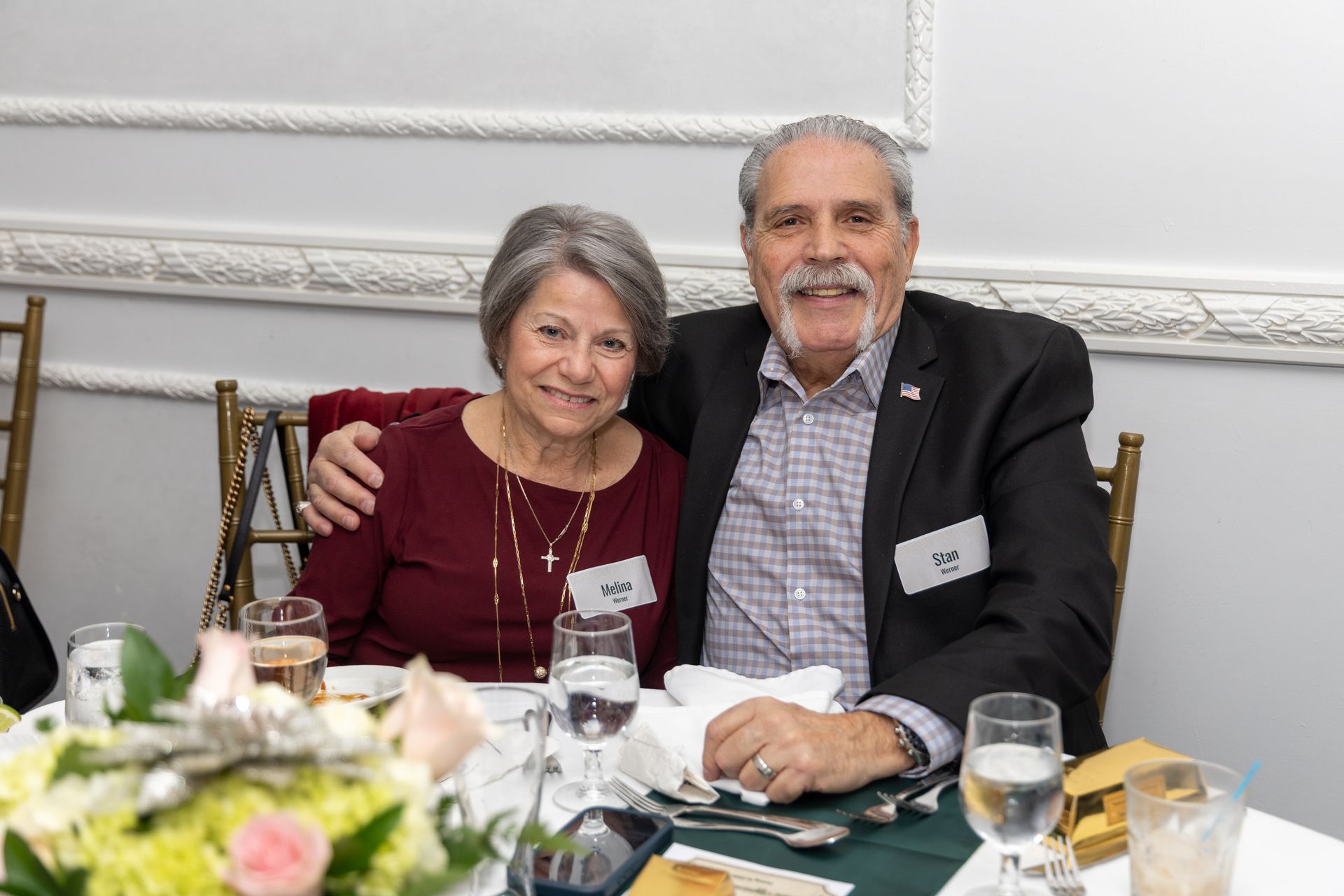 Couple smiling at a table, man with arm around woman. Formal setting, glassware and flowers.