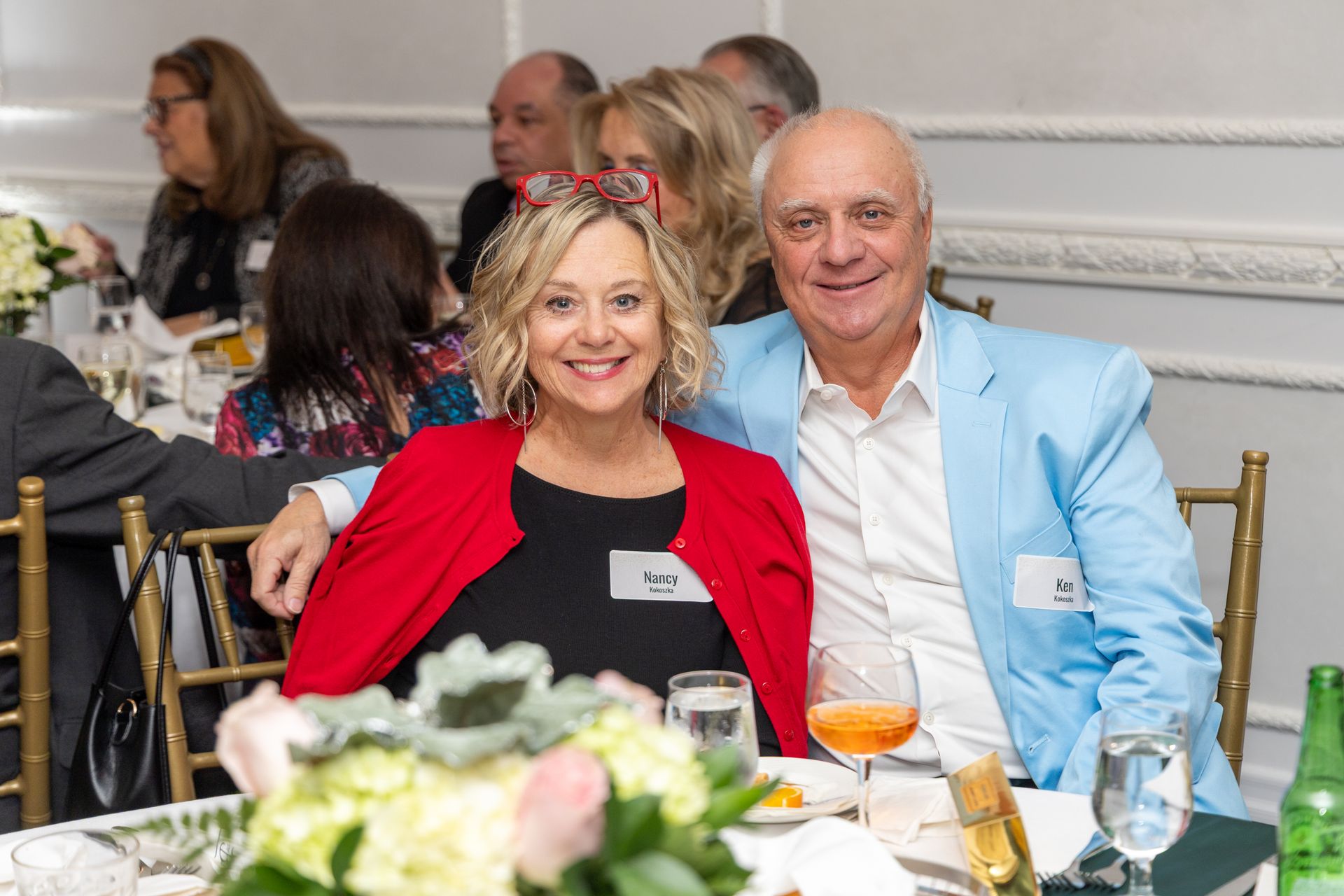 Smiling couple at a formal dinner, woman in red jacket, man in blue blazer, seated at a decorated table.