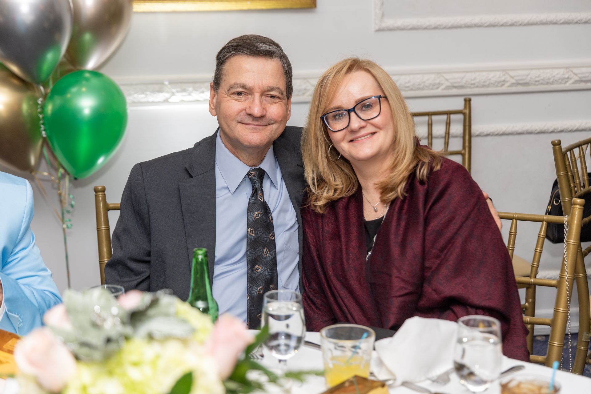 Man and woman smiling at a table, likely at an event. Balloons and floral arrangement in the background.