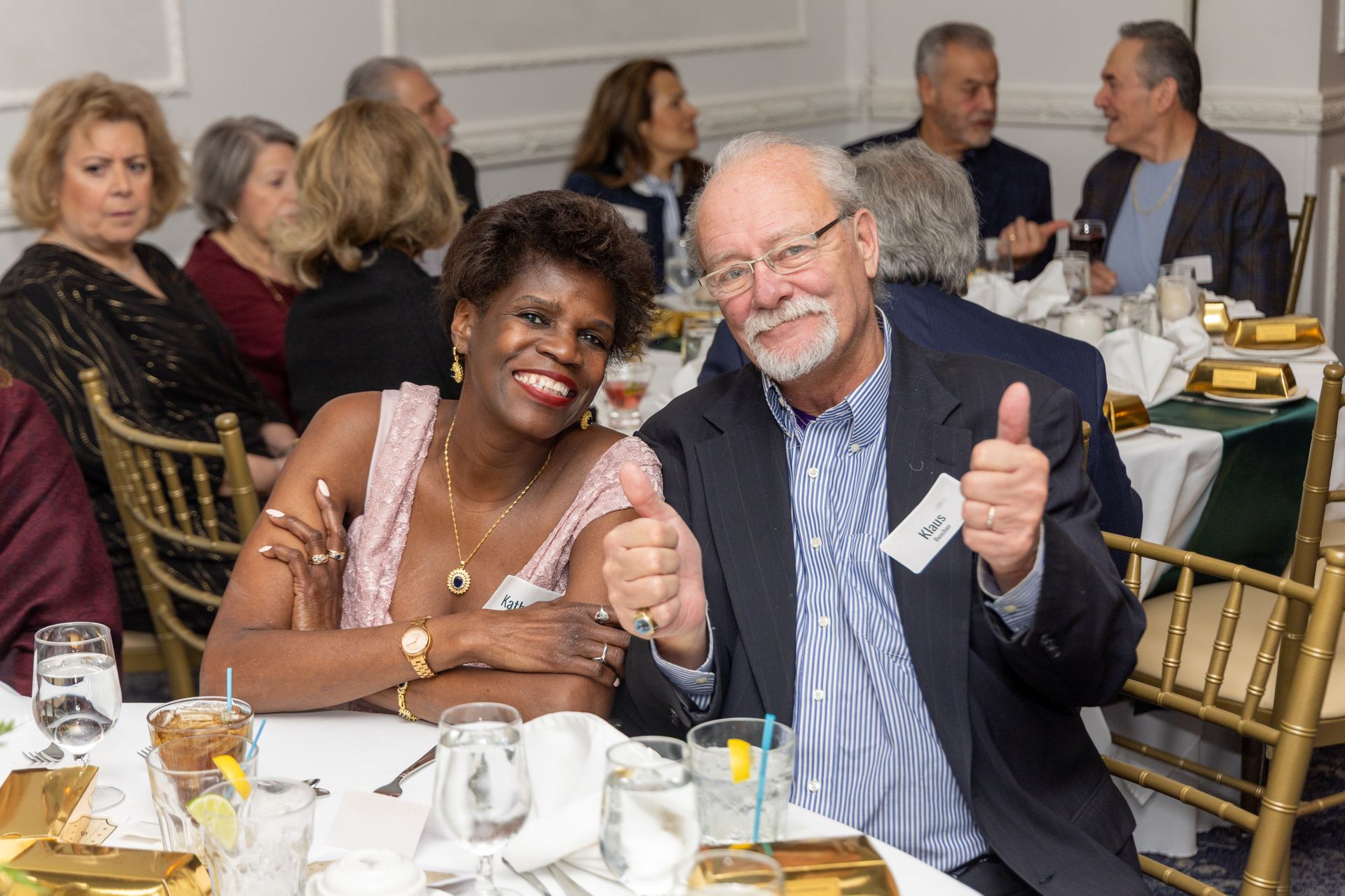 Two people smiling and giving thumbs up at a table; others seated nearby in a well-lit room.