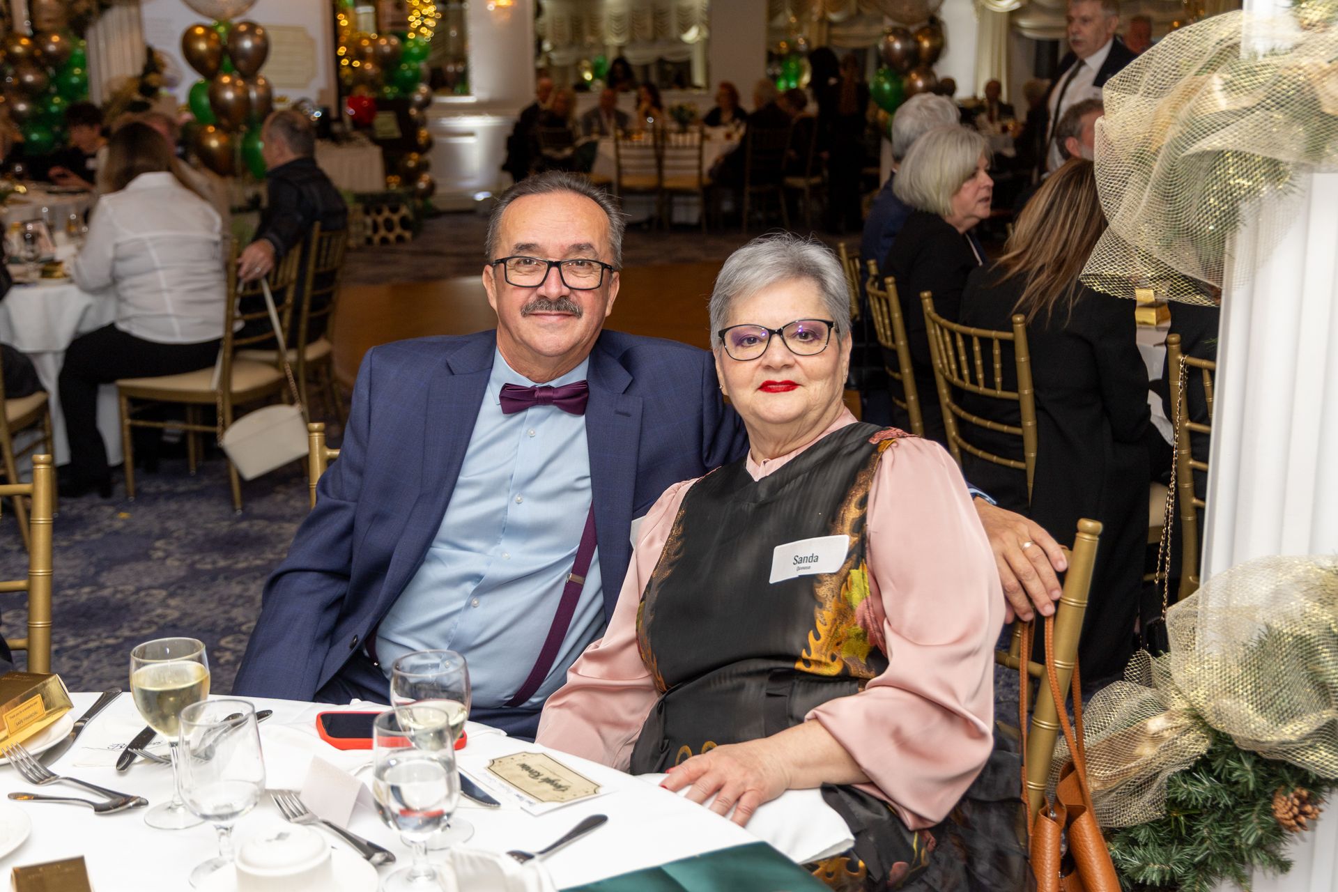 Couple seated at a decorated table, man in blue suit and bow tie, woman in pink blouse, at a formal event.