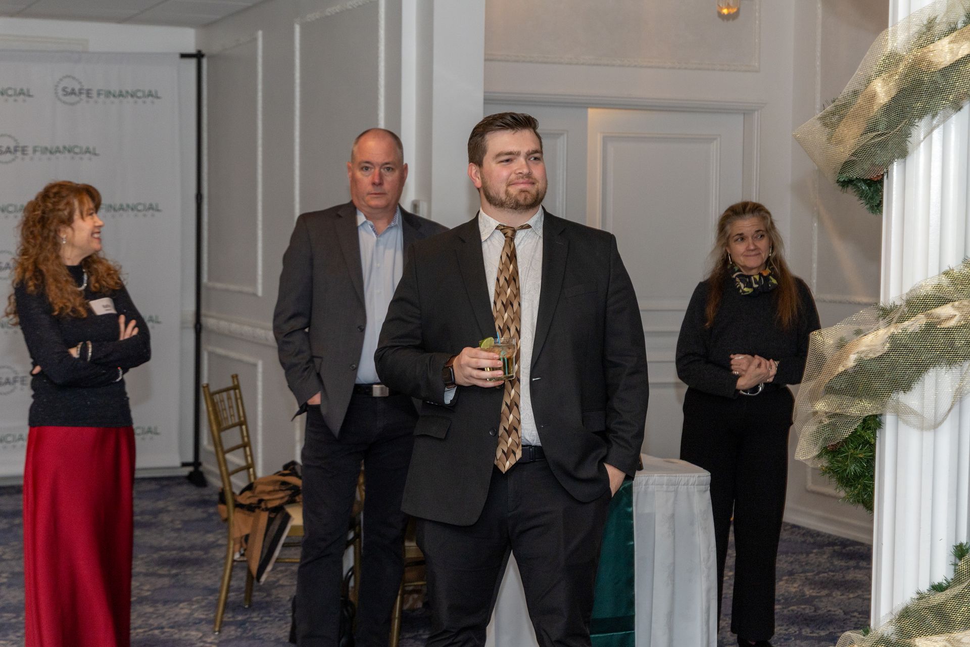 Four people in formal attire at an event. Man in suit holds a drink. Others observe. White walls, gold chair.