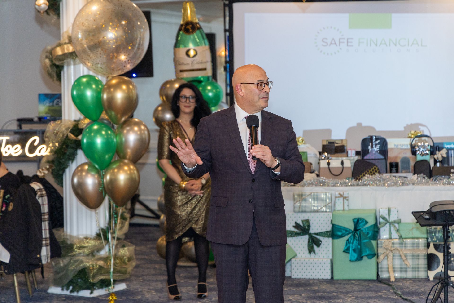 Man in suit speaks at event; woman in gold dress stands behind him near balloons and gifts.
