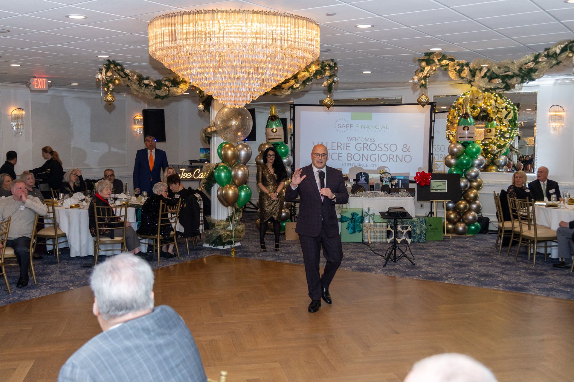 Man speaking at an event with gold and green decorations, a chandelier, and an audience at tables.