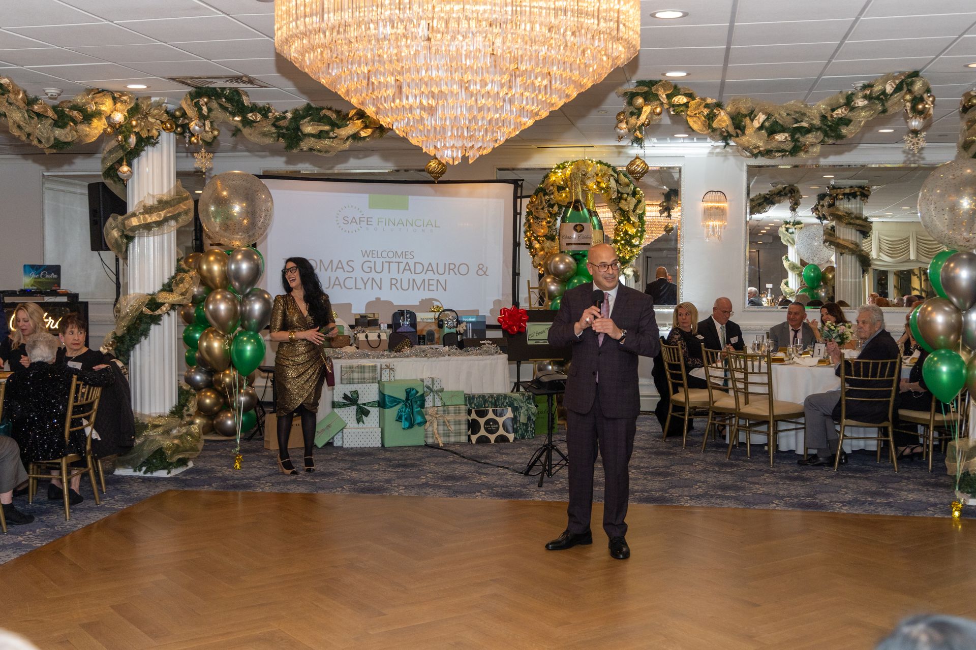 Man speaking at a gala. Woman stands nearby. Room decorated with balloons and garland. Guests at tables.