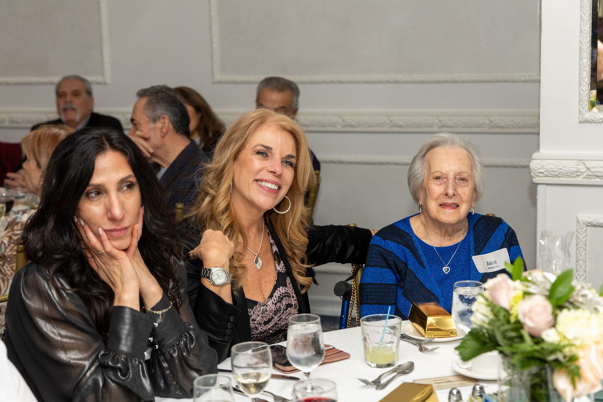 Three women seated at a table, smiling, at an event. White tablecloth, flowers, people in background.