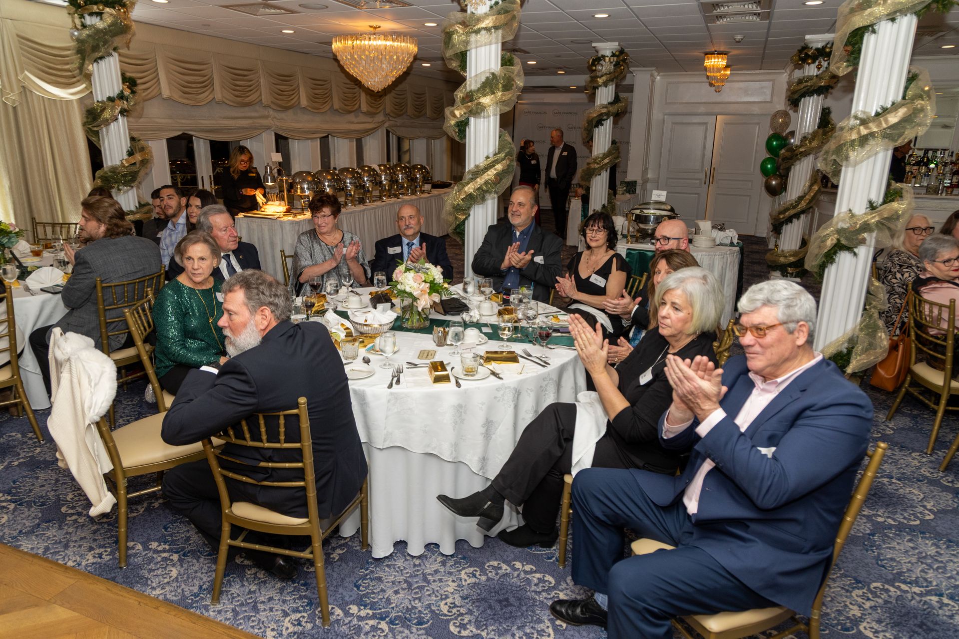 People seated at tables in a decorated room, clapping. Gold and green decorations, a chandelier, and a buffet are visible.