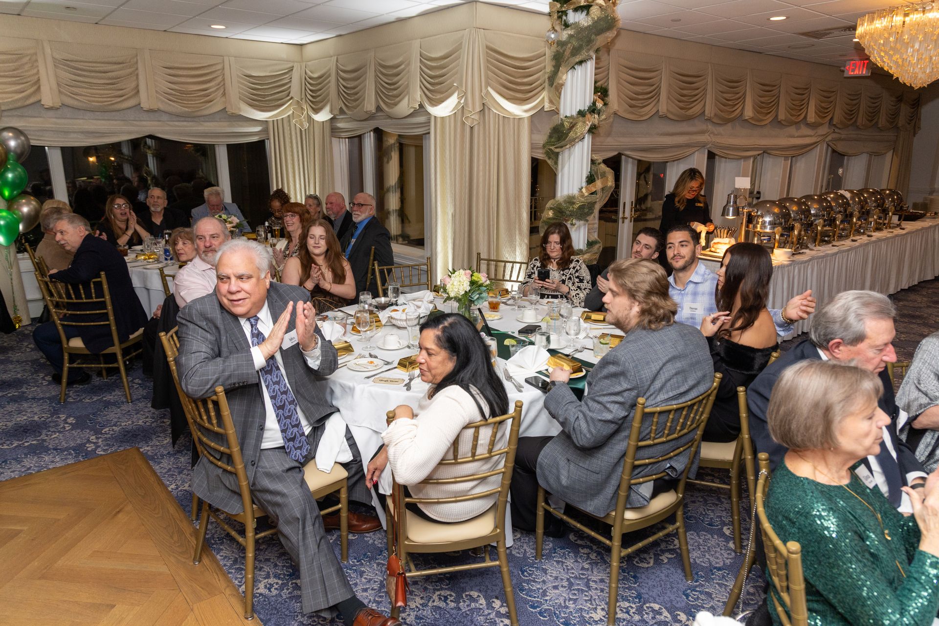 People at a formal event, seated at round tables with centerpieces. Elegant room, buffet in the background.