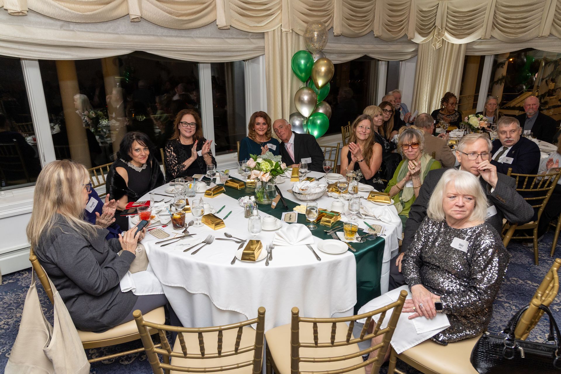 People at a formal event seated around a decorated table with centerpieces. Balloons in the background.