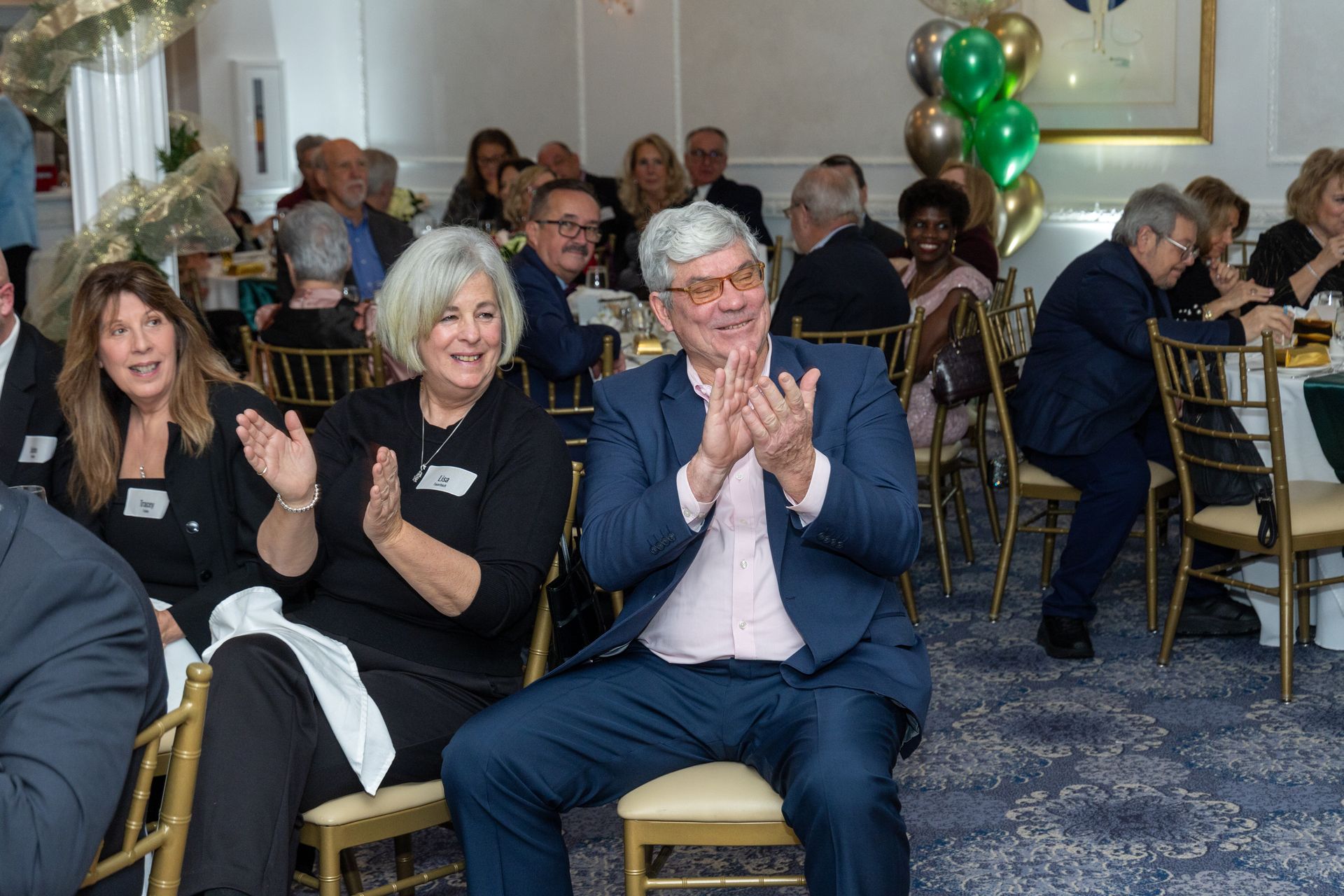 People applauding at a formal event, seated at a round table. Balloons and decorations visible.