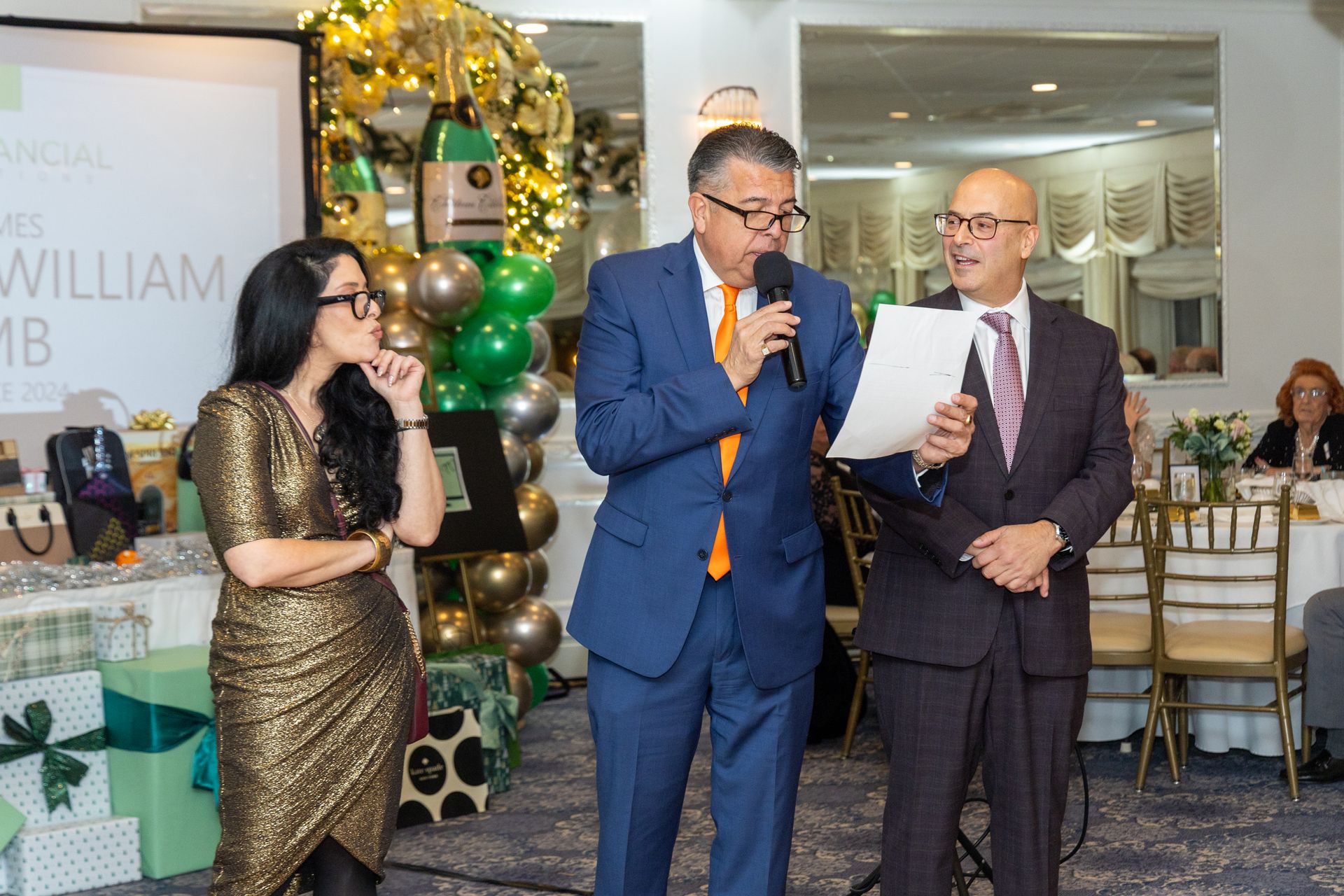 Three people at a formal event; a man speaks into a microphone, another reads paper, a woman observes, green and gold decor.