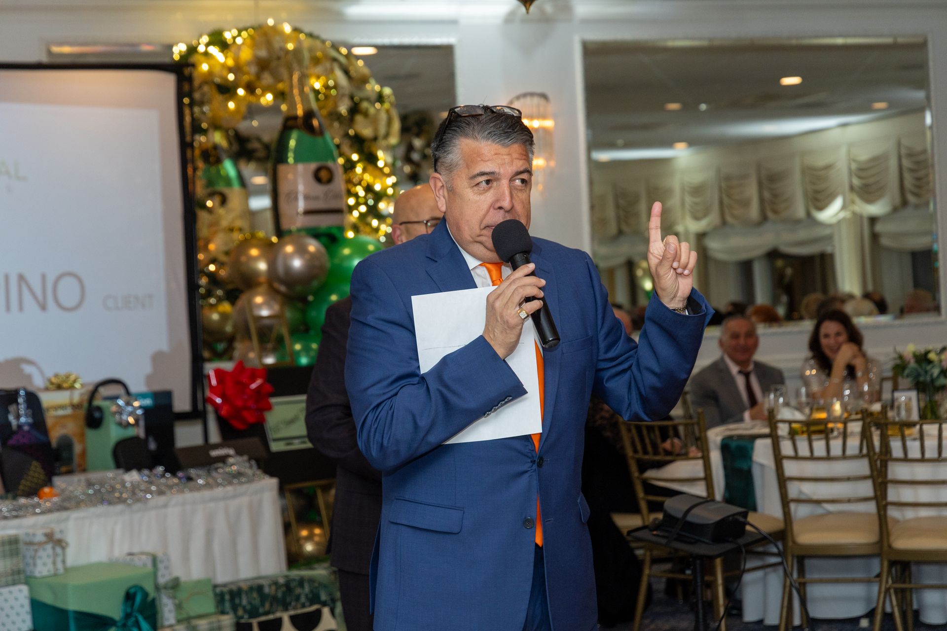 Man in blue suit speaking into microphone, gesturing with one finger, at an event.