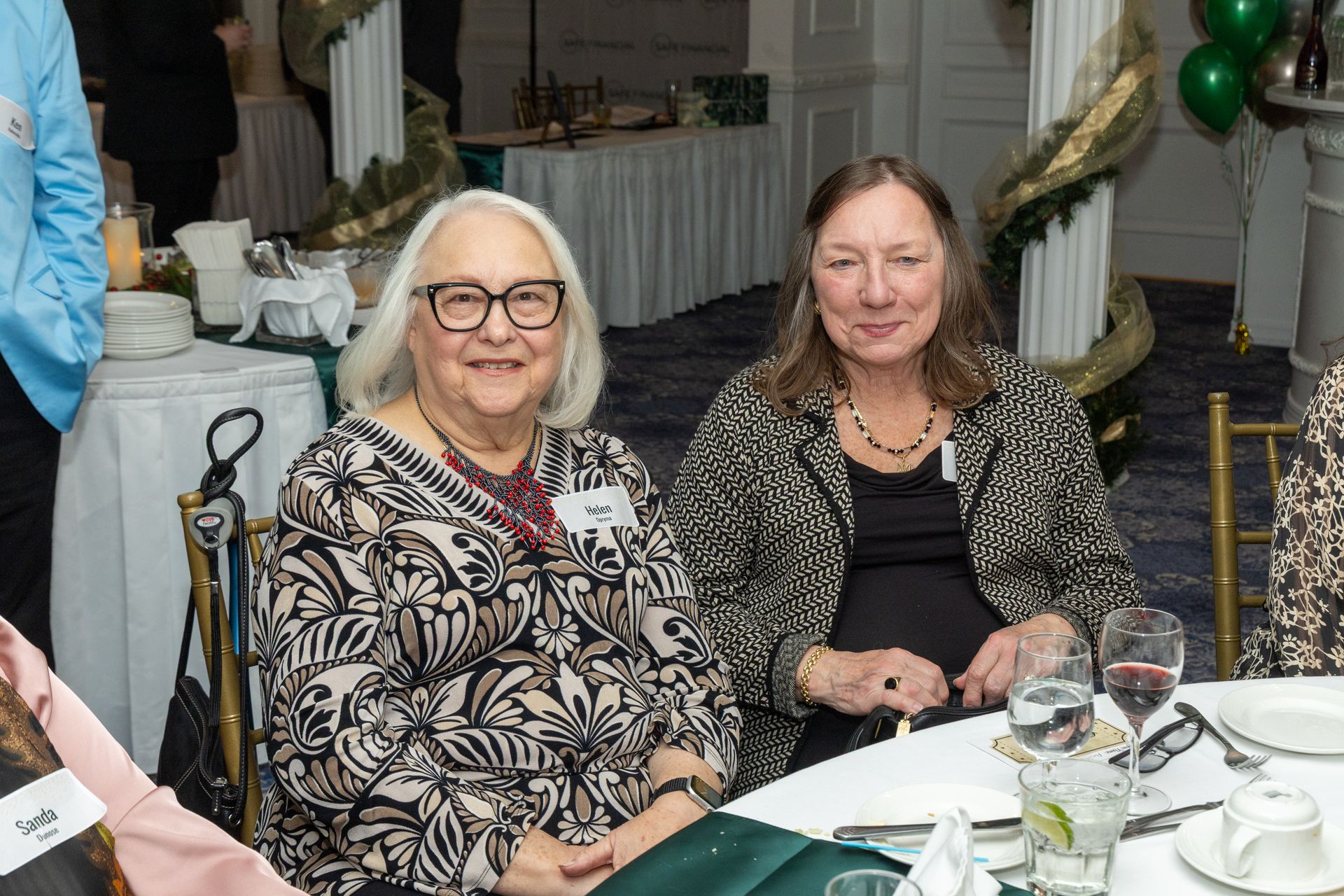 Two women smiling at a table, at an event. One wears glasses, a patterned shirt. Other wears jacket.