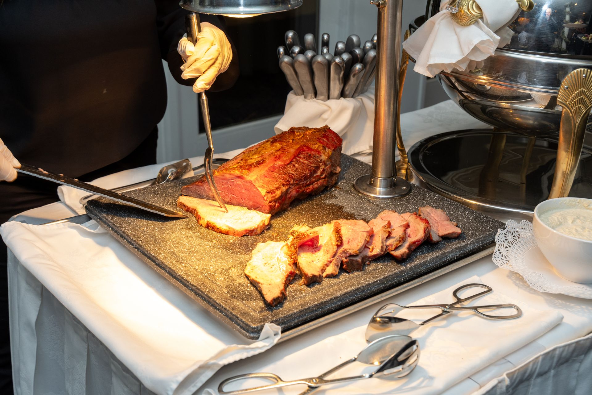 Roast beef carving station at an event with sliced meat, tongs, and a server.