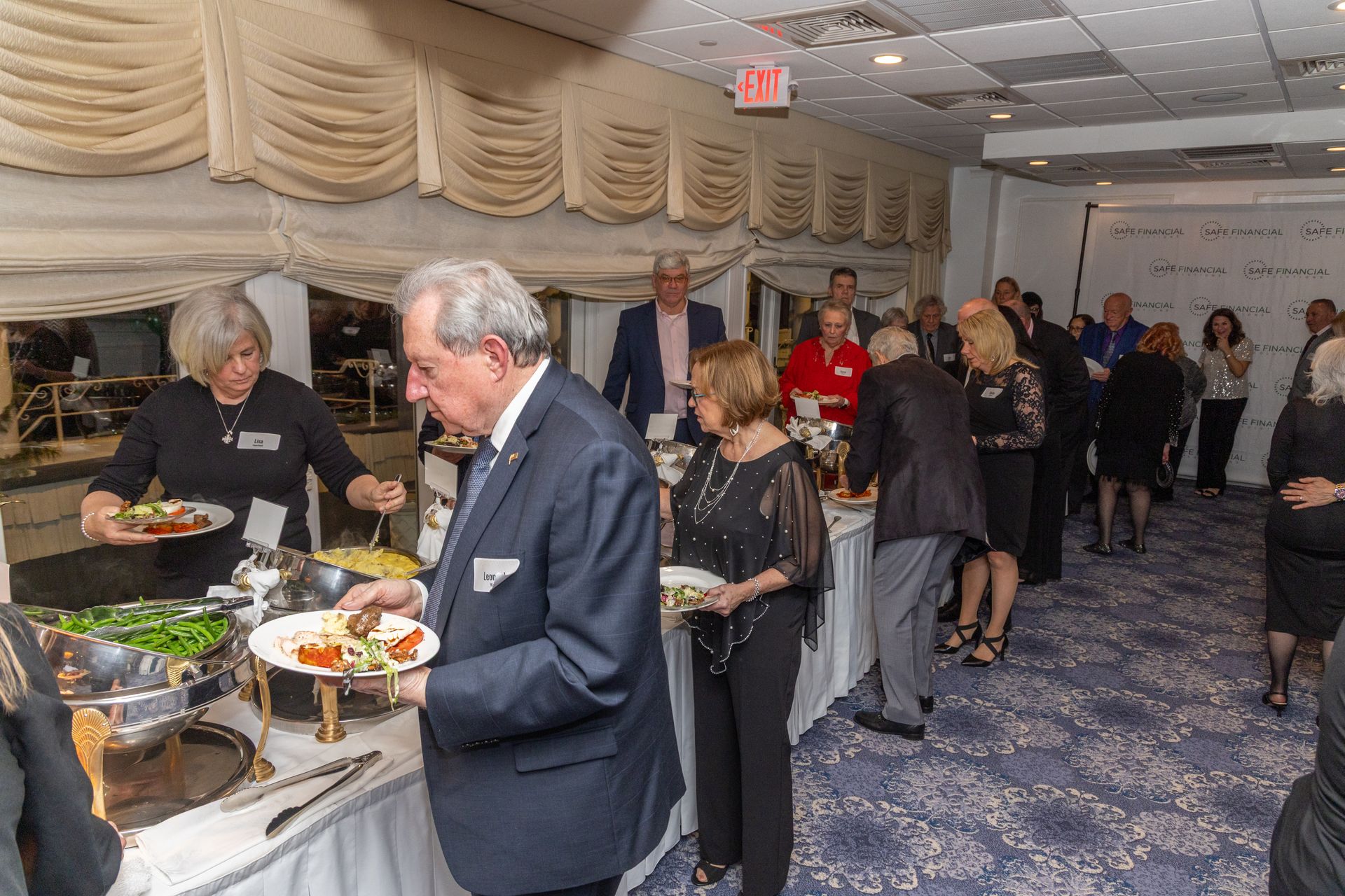 People at a buffet, serving food. Indoors, many guests and serving staff, white tablecloths, and blue carpet.