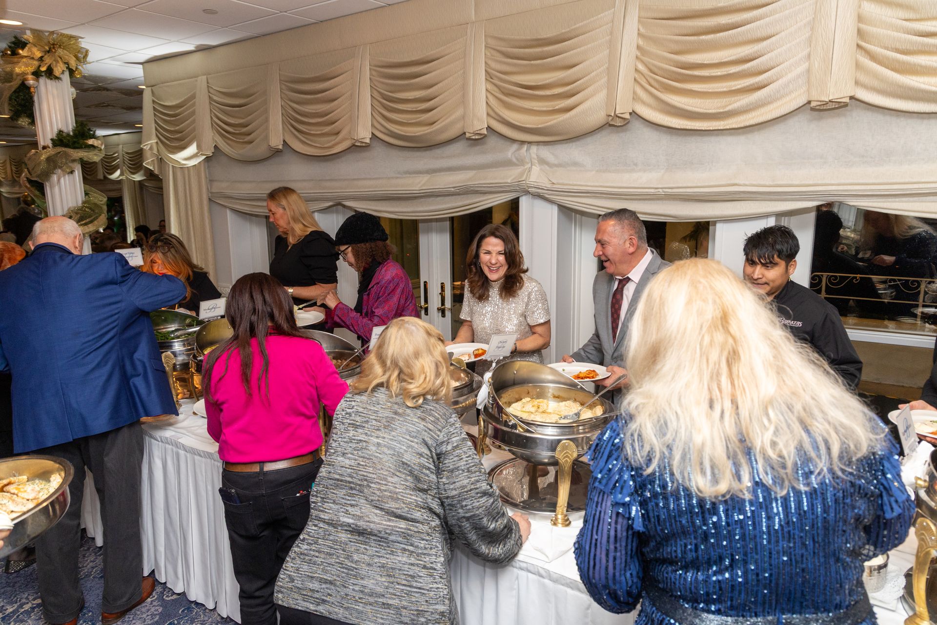 People at a buffet table, serving food from warmers in a well-lit room, possibly at a party or event.