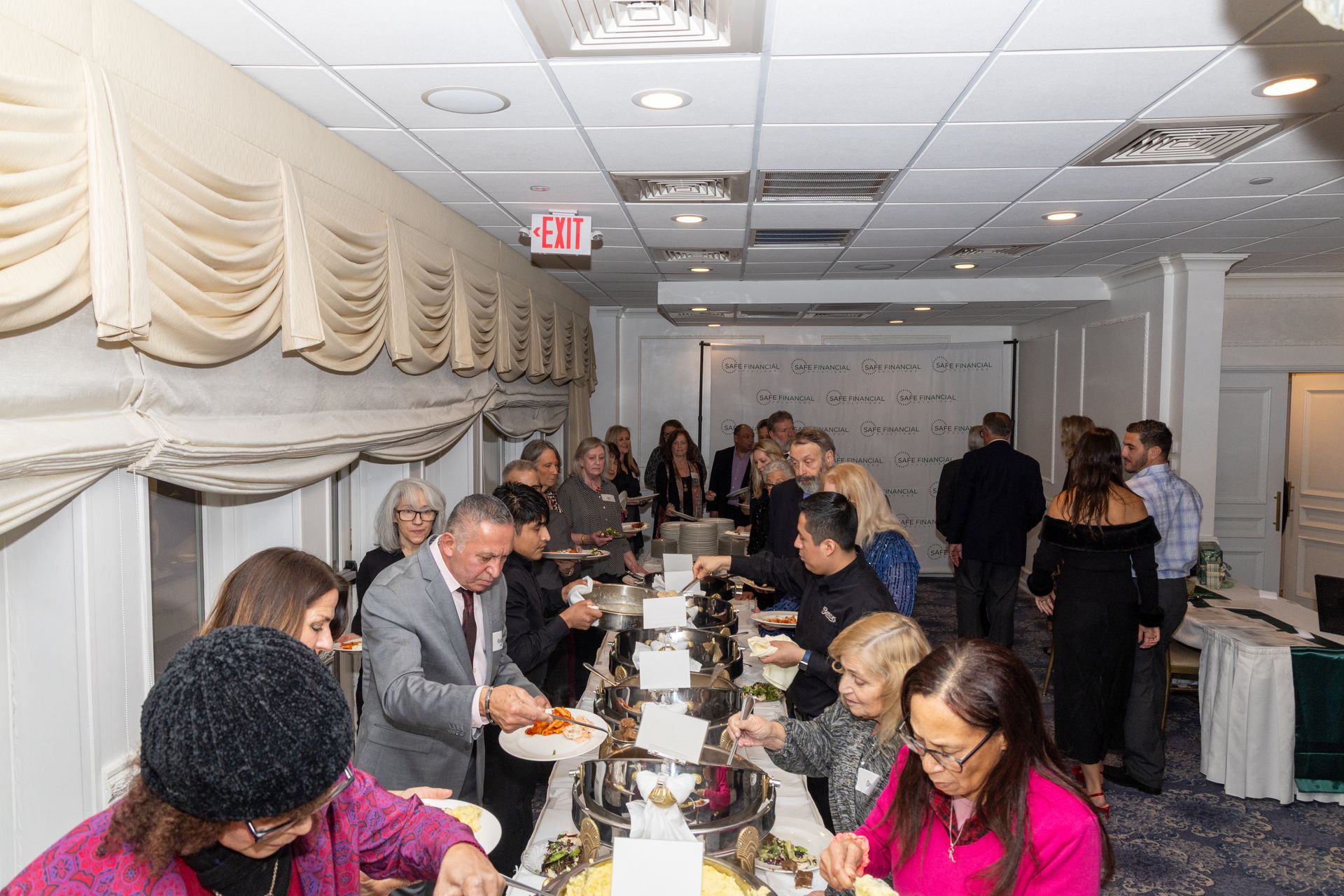 People at a buffet table in a banquet hall, selecting food from various silver serving dishes.