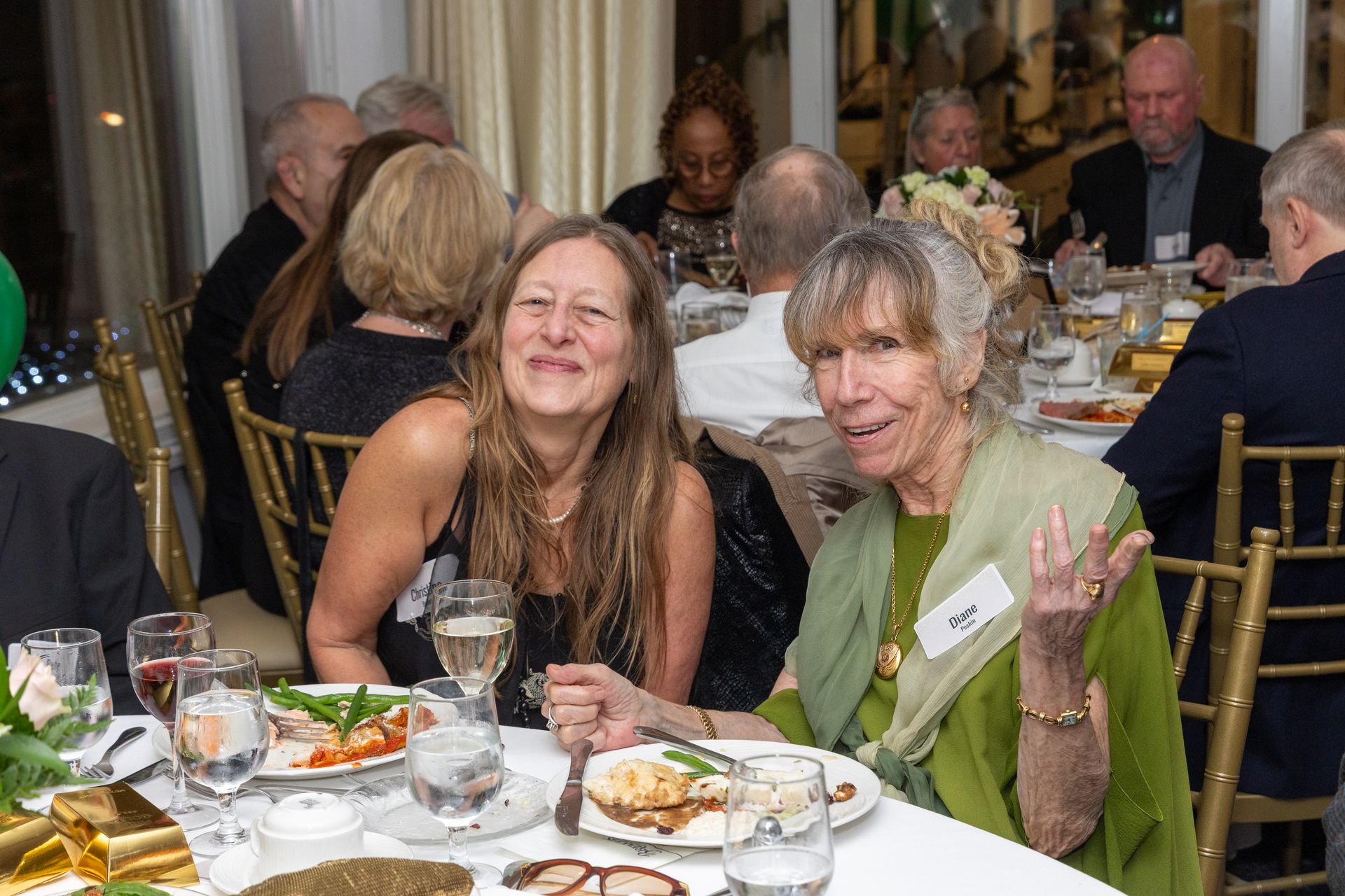 Two smiling women at a dinner table, with others in the background. One waves her hand.