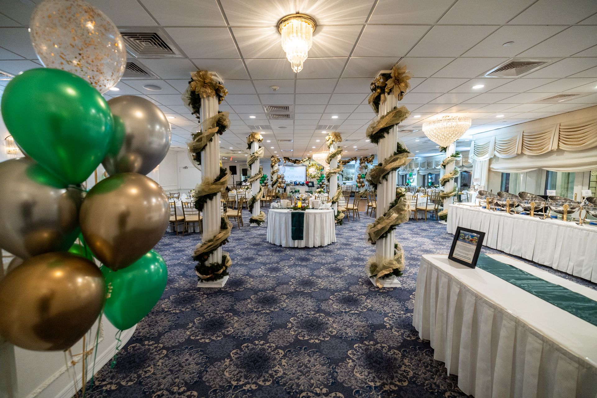 Party venue with decorated columns, tables, and balloons in green, gold, and silver.