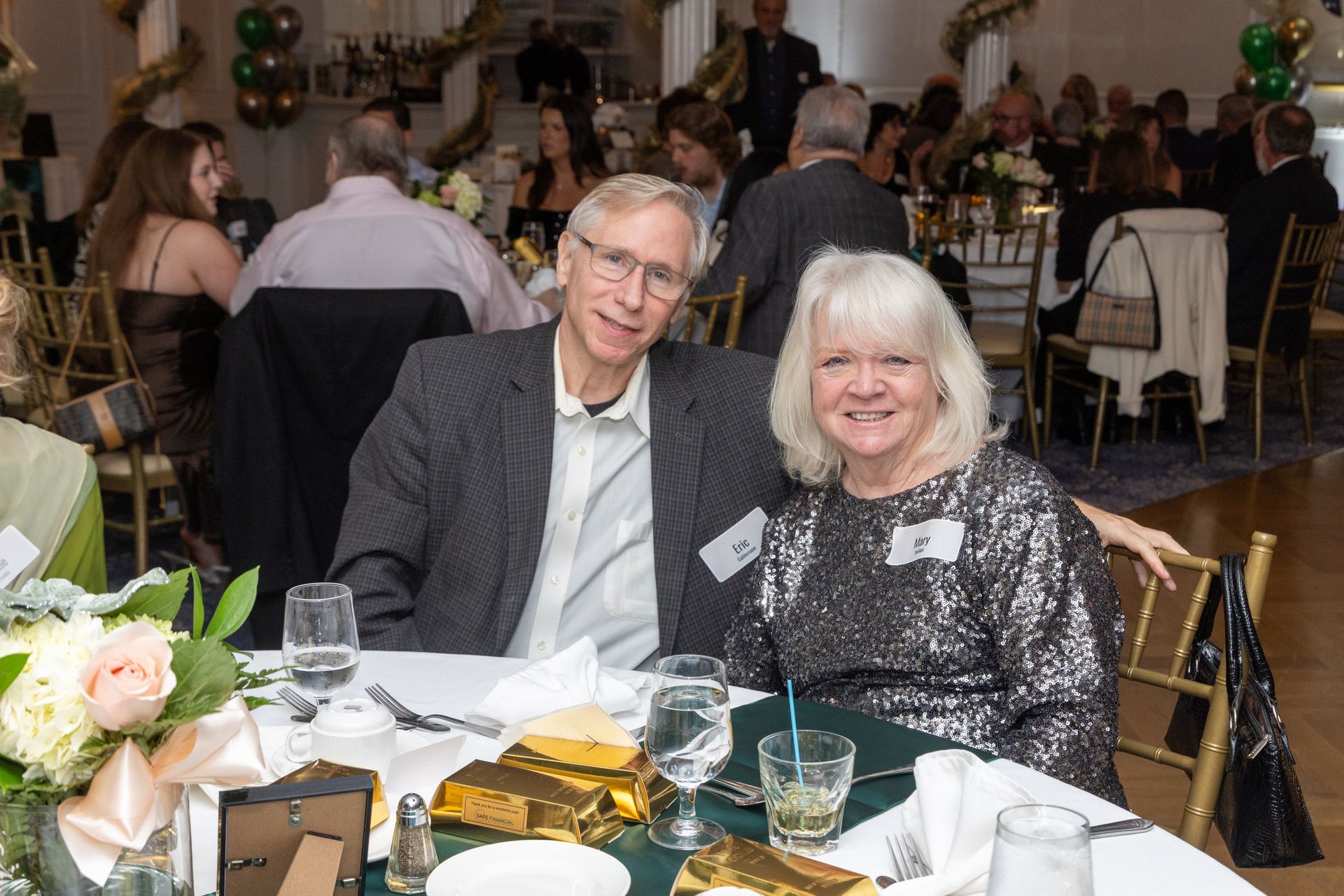 Couple smiling at a table during a formal event. Decorations and other guests are in the background.