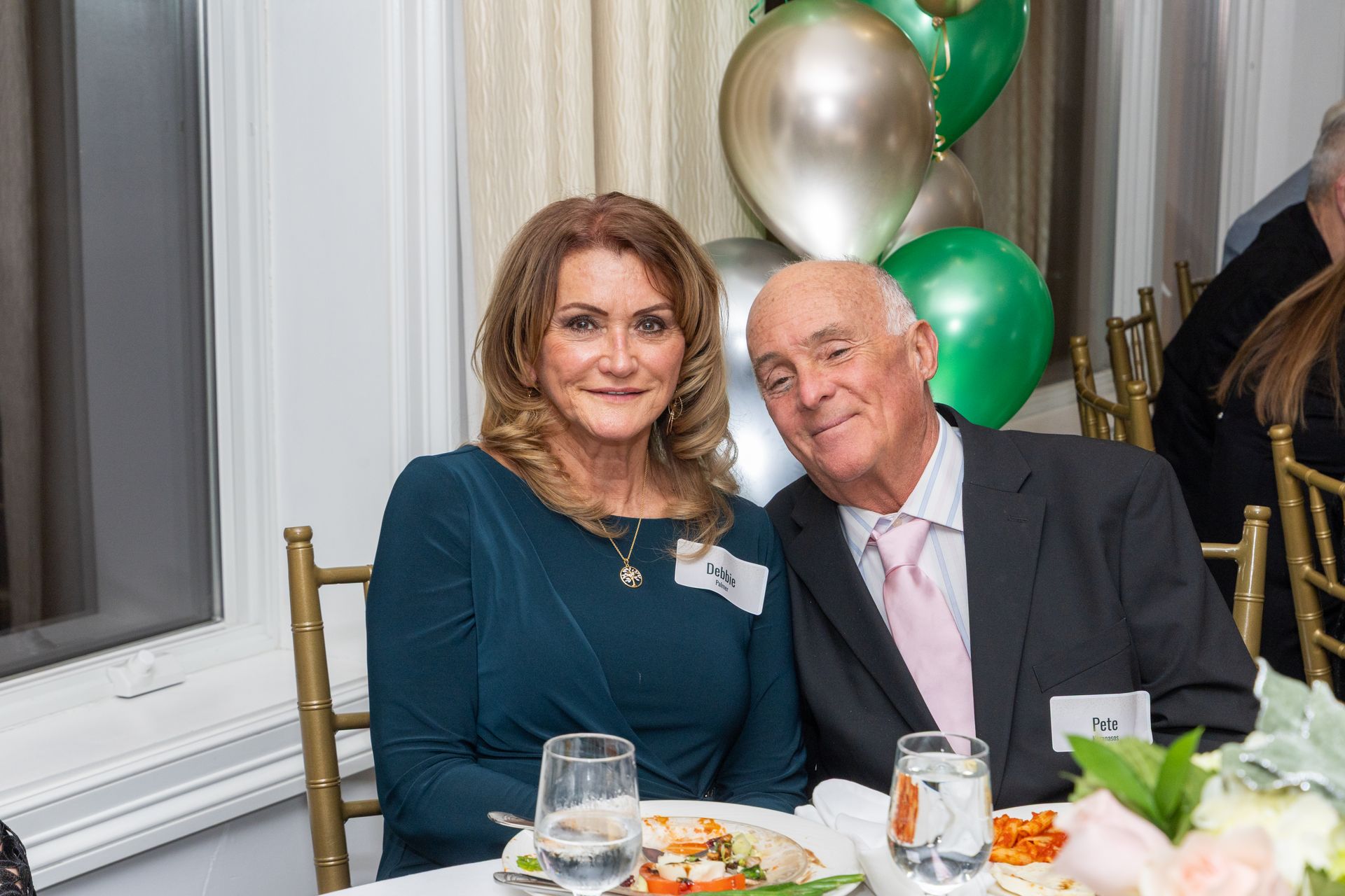 A woman and man smiling at a table with food, balloons in the background.