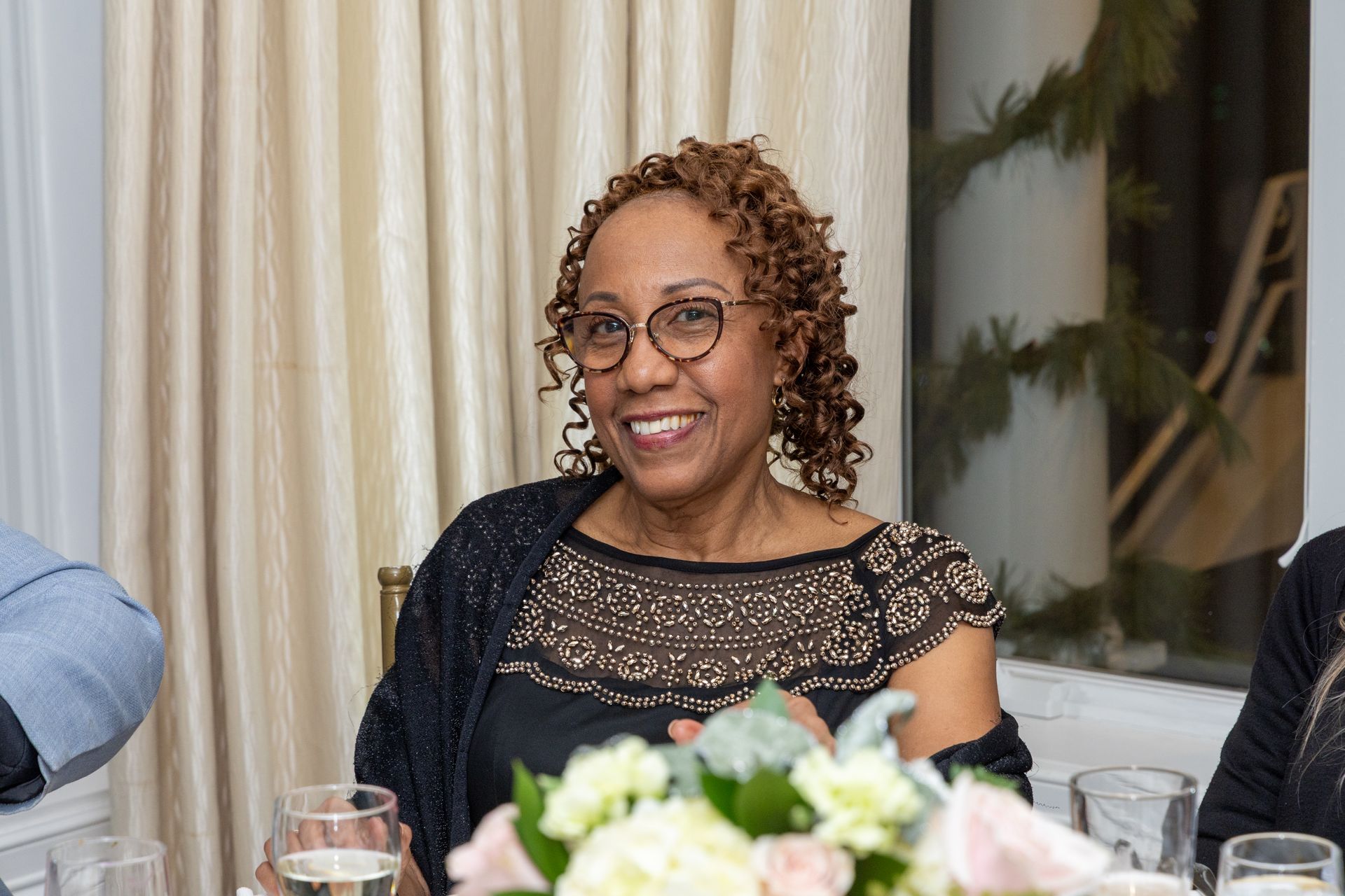 Woman with glasses smiles at a table with flowers. She wears a black top and a shawl, inside.