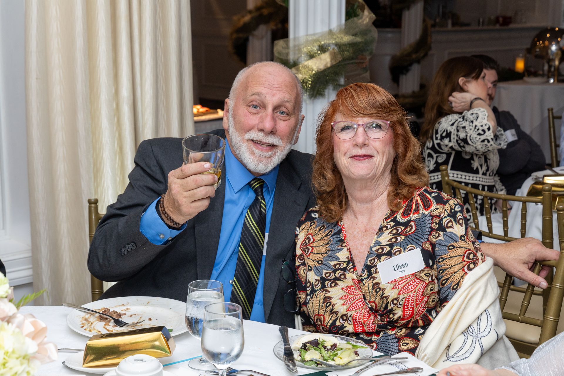 Man in suit raises glass, smiling, beside woman in floral shirt at table.