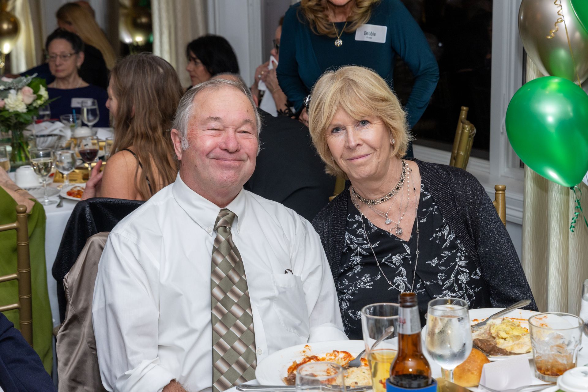 Couple at a dinner table smiling, indoors; green balloons in background.