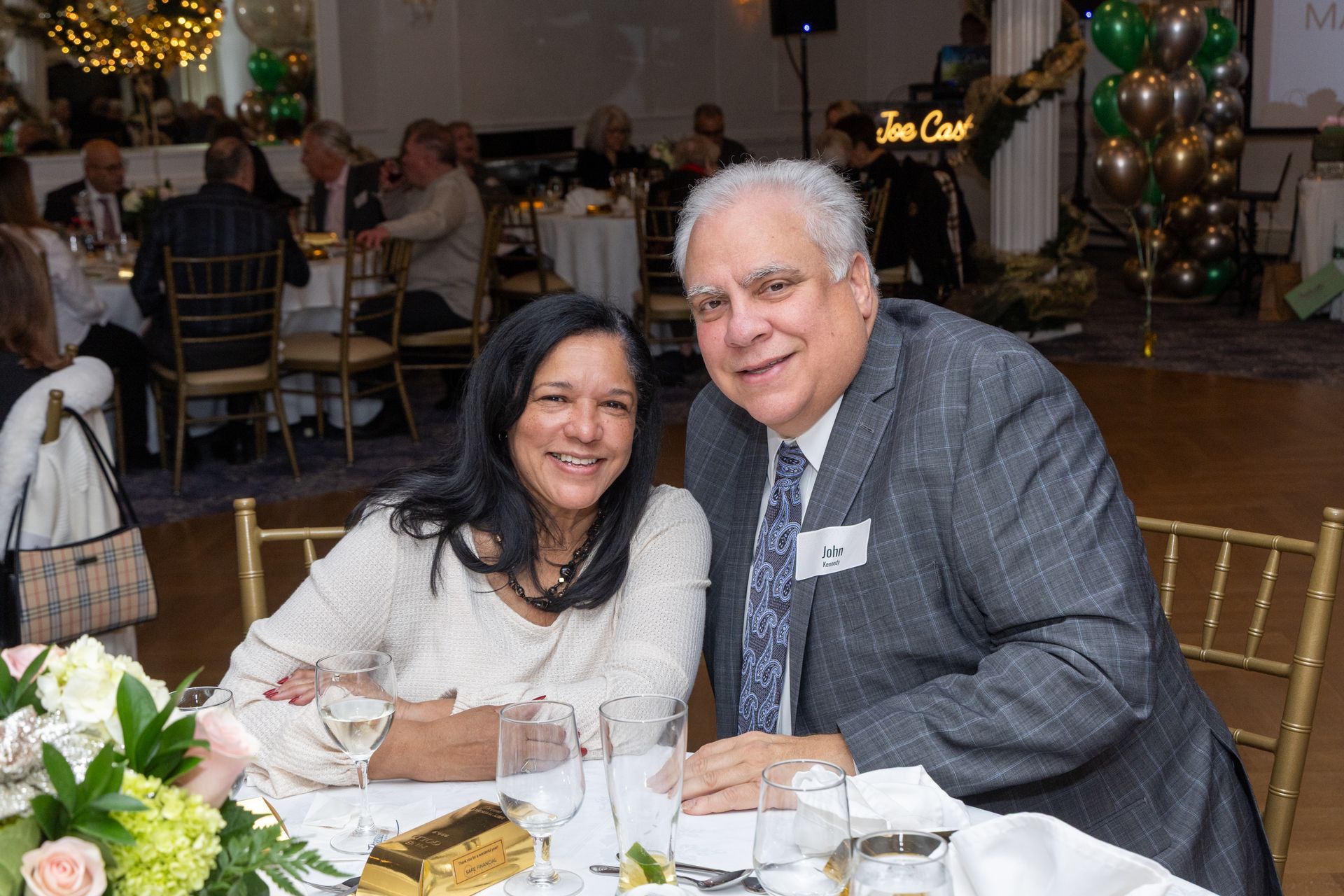 Couple smiling at a formal event, seated at a table. Guests and decorations visible in the background.
