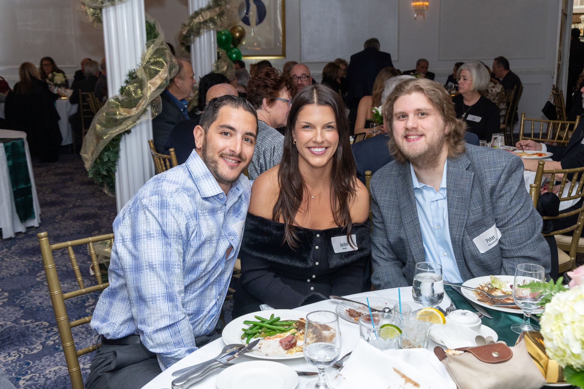 Three people smiling at a table; one woman flanked by two men. Formal event with round tables and decorations.