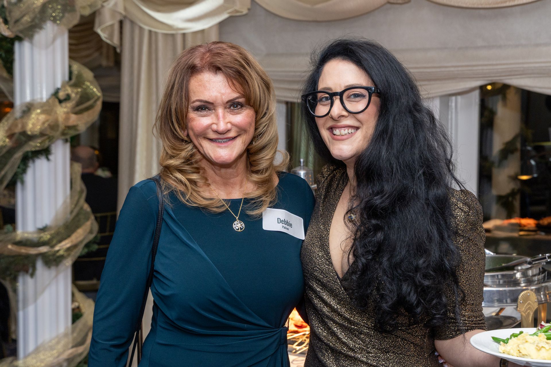 Two women smile at the camera. One wears a teal dress, the other a gold top, black glasses, and long dark hair.