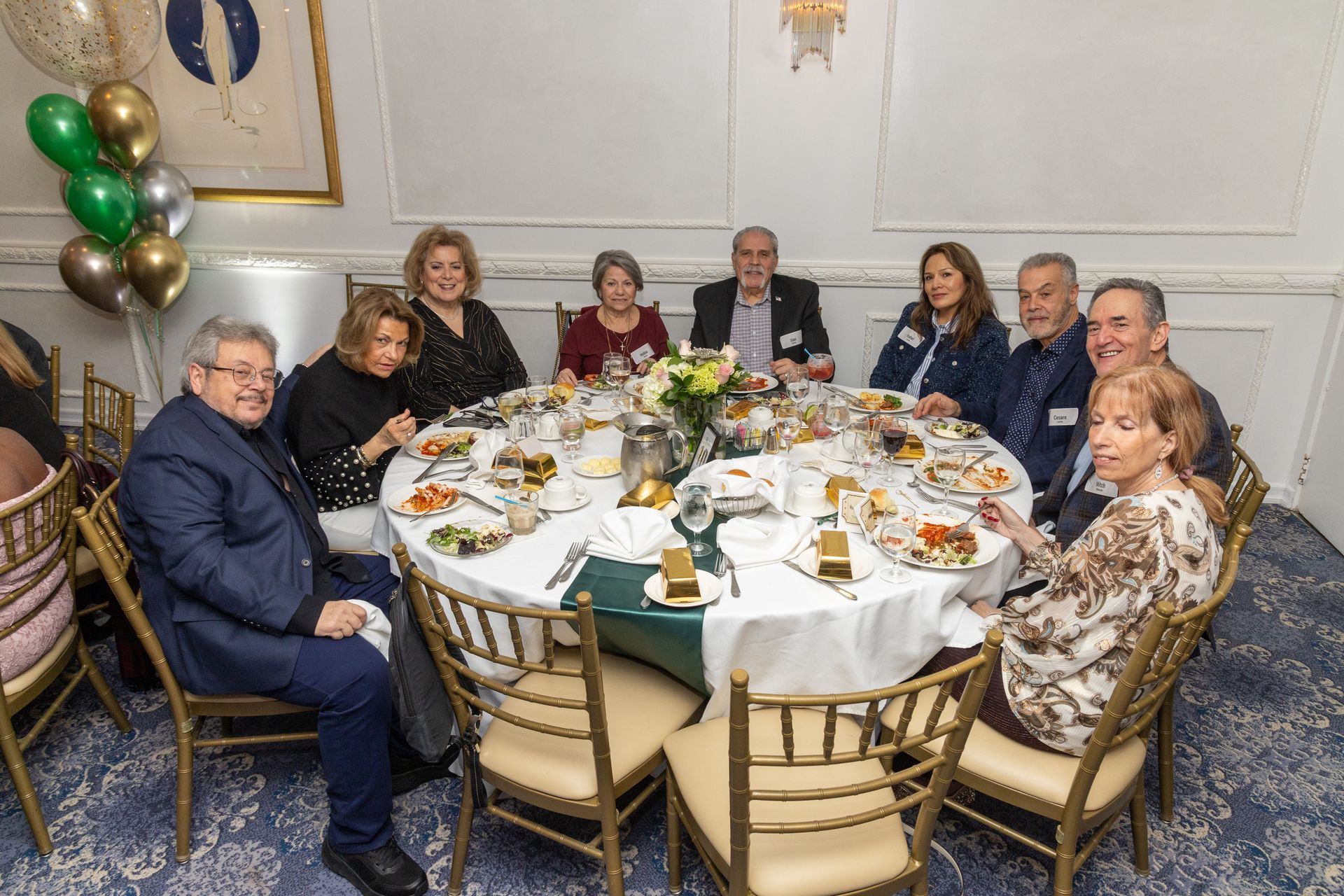 People seated around a round dining table. Food and drinks are present. Balloons decorate the room.
