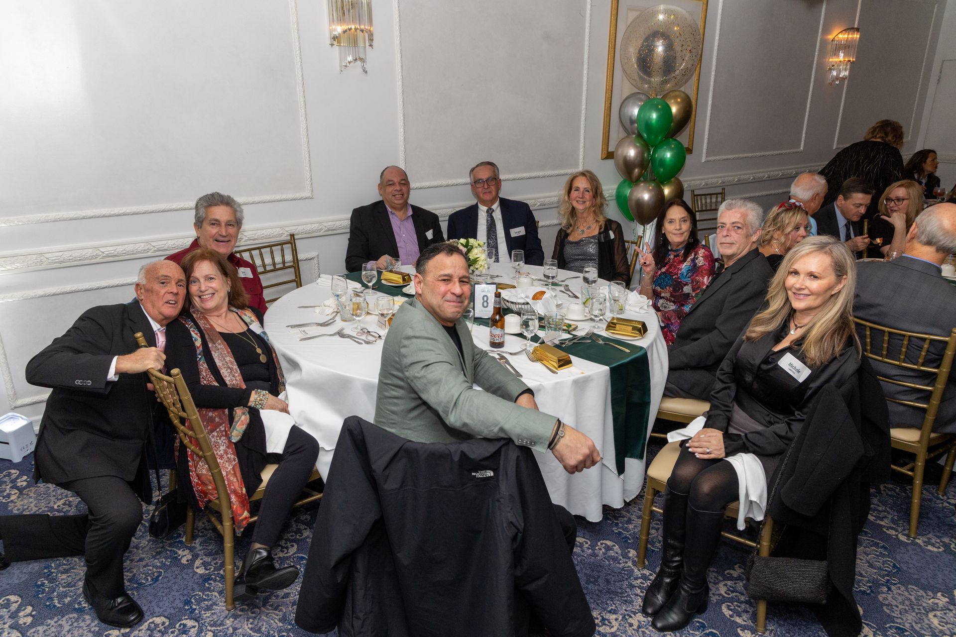 People seated at a round table, celebrating. Green and gold balloons. Formal attire. Indoors.