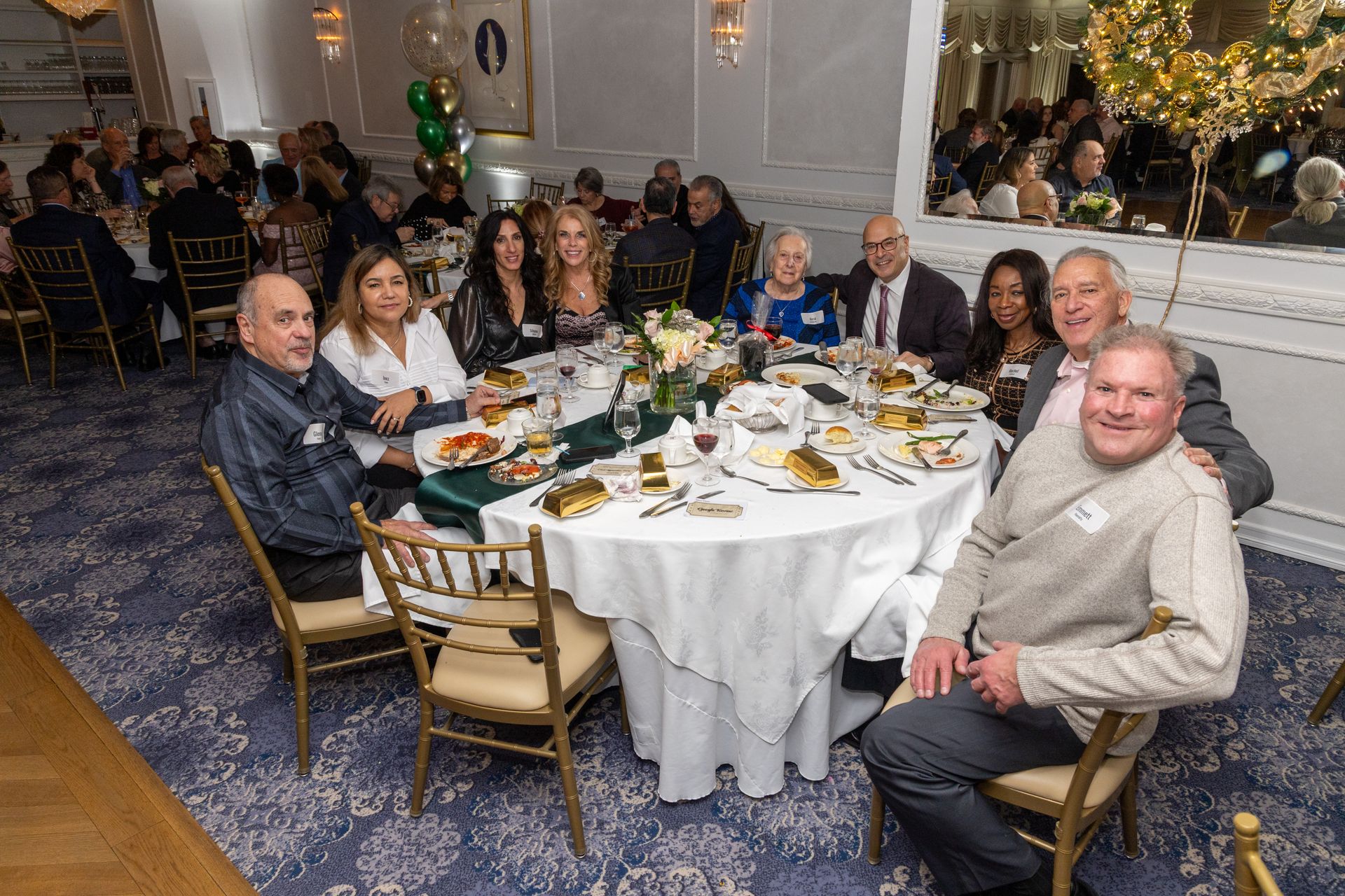 People seated at a round table, indoors, celebratory event, white tablecloth, gold chairs, looking at the camera.