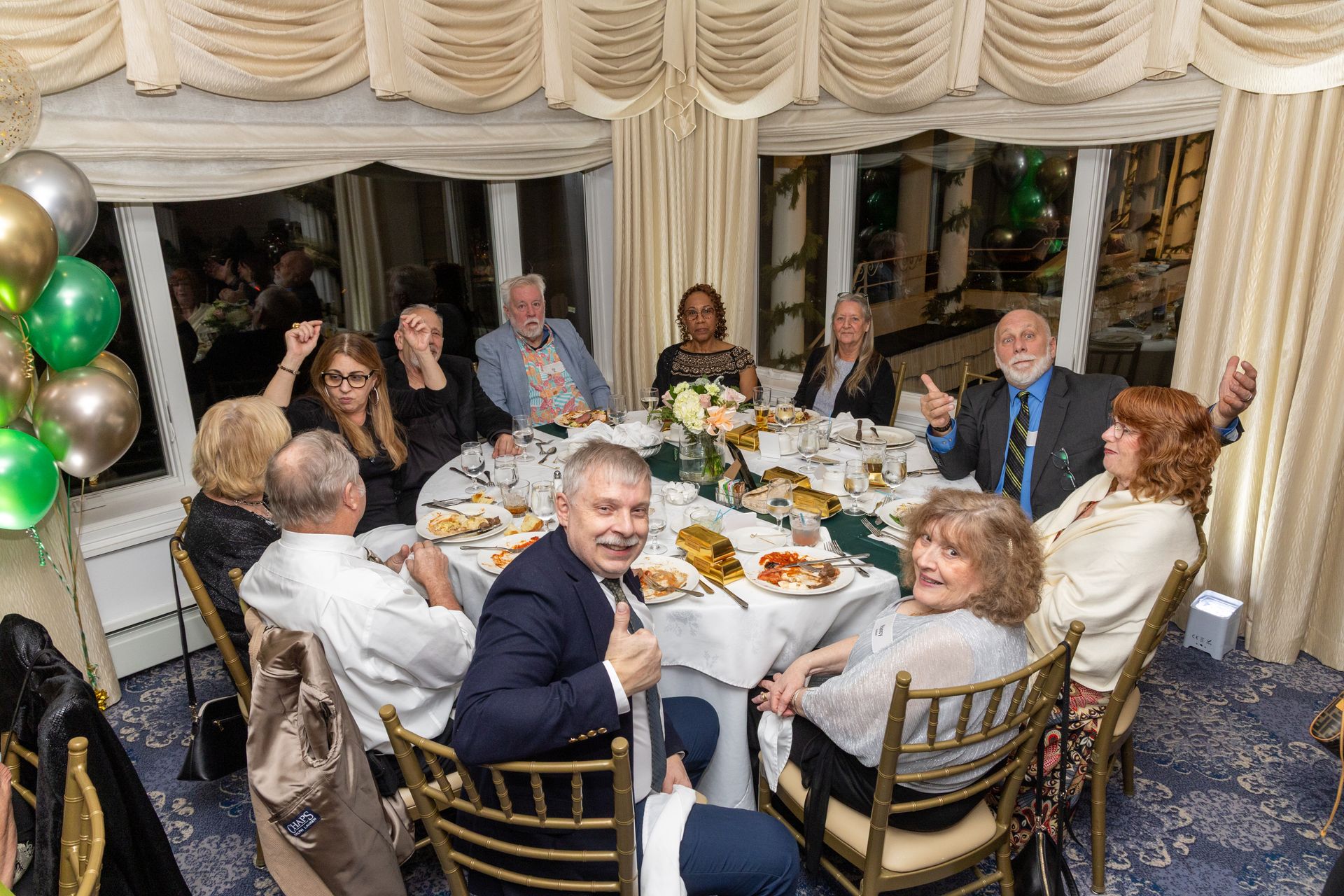 Group of people seated around a round table in a dining room, celebrating with food and balloons.