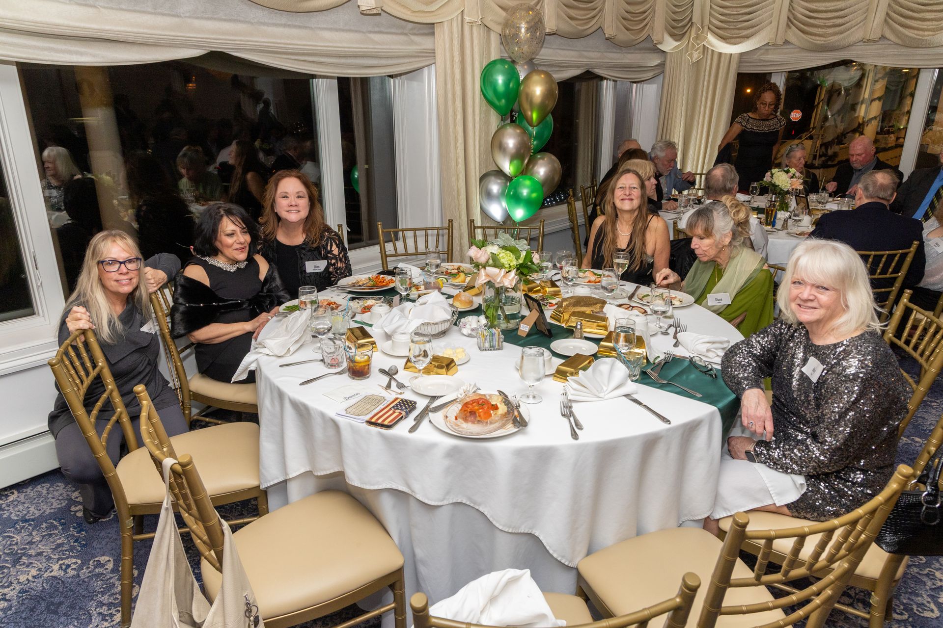 People seated around a decorated round table in a banquet hall. Balloons in the background.